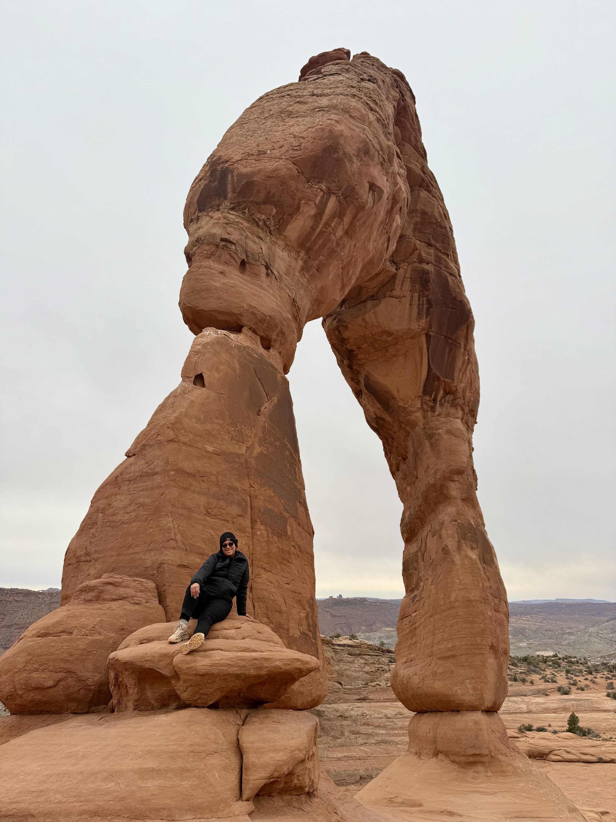 A woman relaxing on a large rock at the base of Delicate Arch in Arches National Park, Utah, with the arch towering above against a cloudy sky.