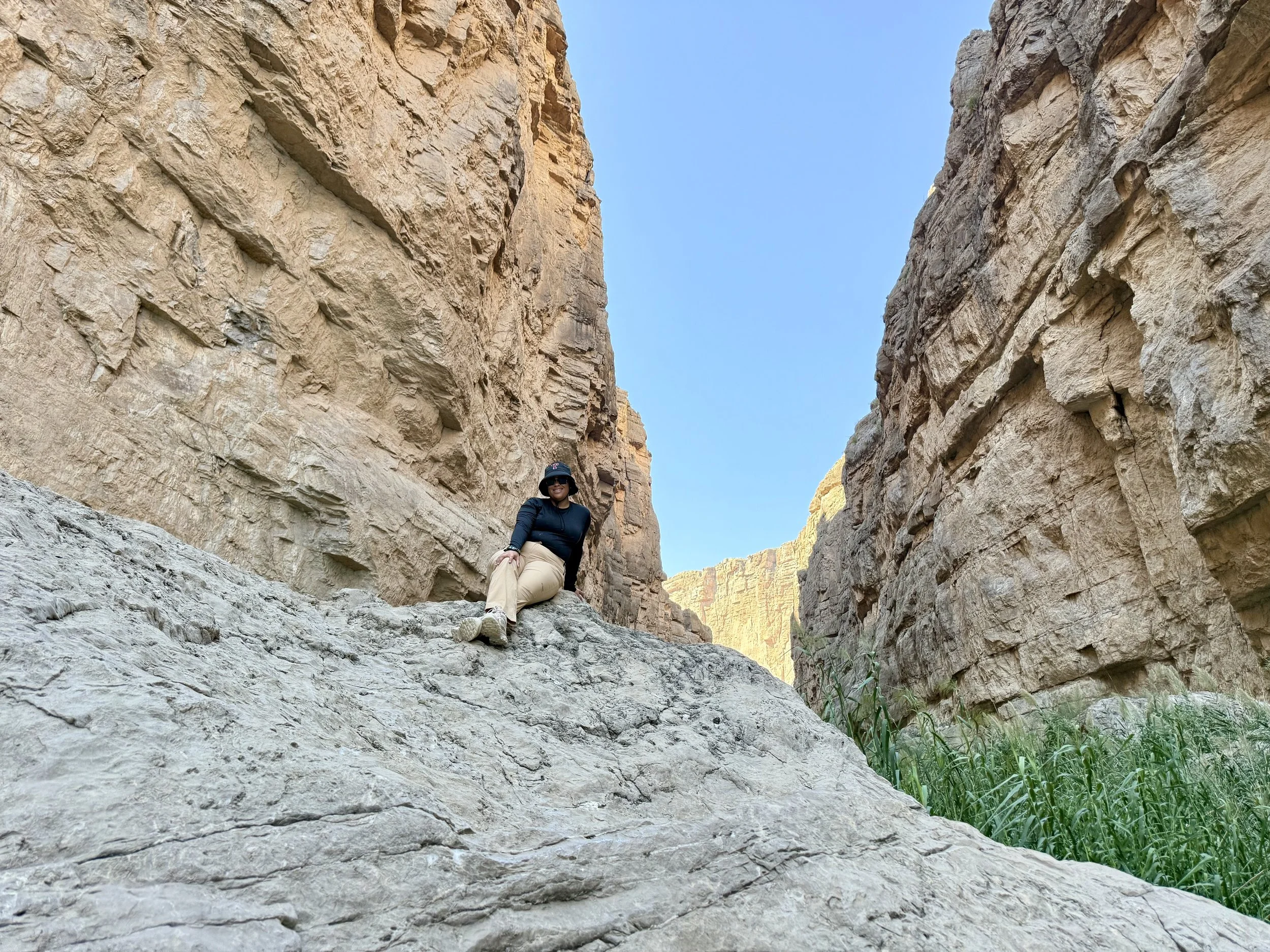 Woman sitting on a large rock in a canyon with tall beige rock walls and green plants in the foreground, under a clear blue sky.