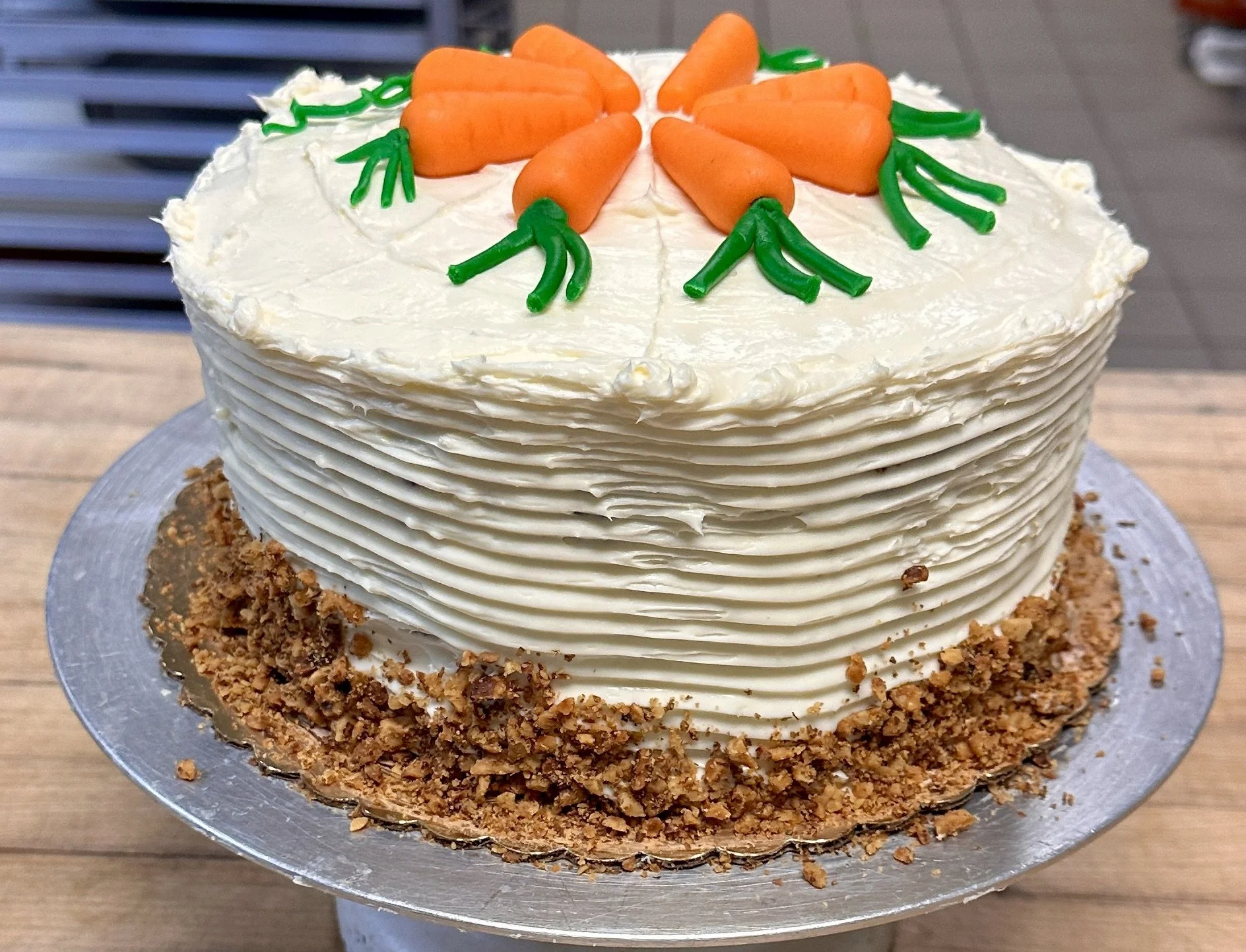 A round carrot-themed cake decorated with orange carrots and green leaves made from icing, with a cream-colored frosting and a crumbly nut or cookie base, placed on a silver cake stand.