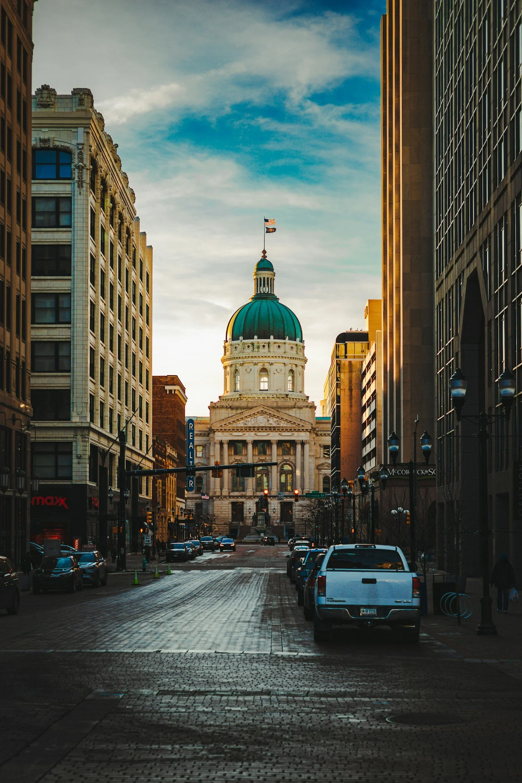 City street leading to a government building with a large green dome and an American flag on top, surrounded by tall buildings at sunset.