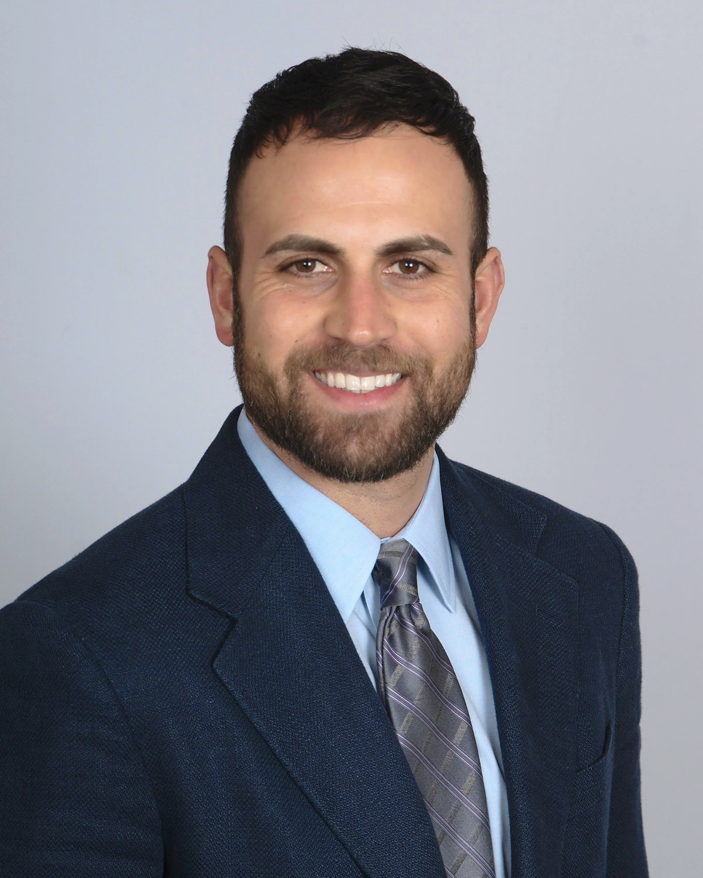 Professional headshot of a man with dark hair and beard wearing a navy suit, light blue shirt, and purple striped tie, smiling against a plain light gray background.