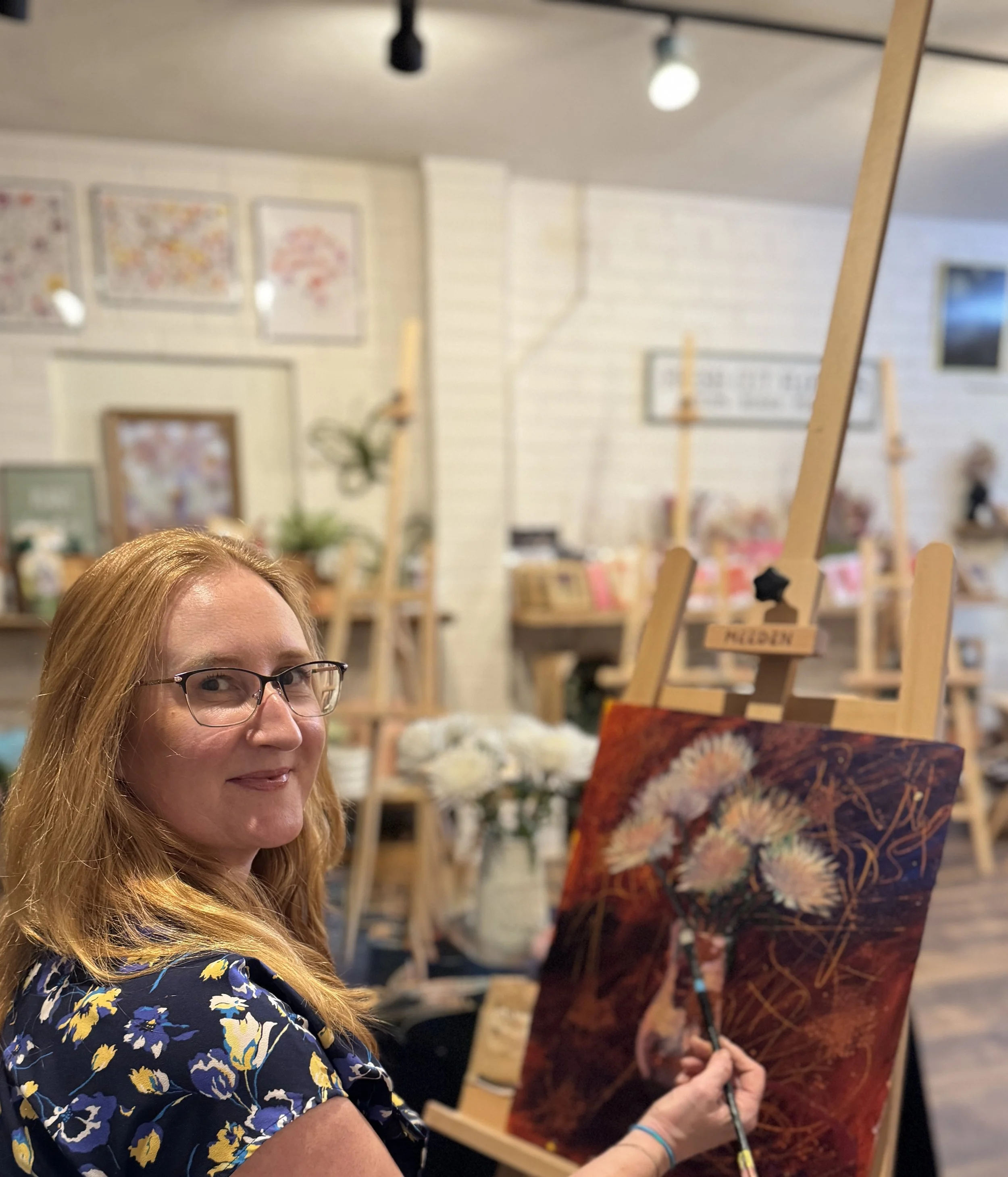 Woman wearing glasses, smiling, painting a floral artwork on a canvas in an art studio with easels and flower arrangements in the background.