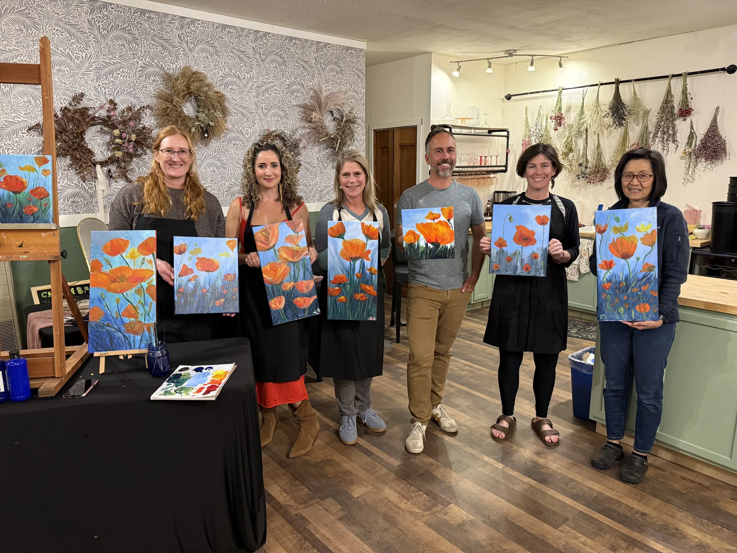 Group of seven people standing in a room, holding paintings of orange poppies. They are smiling and wearing aprons, with art supplies on a table in front of them. The room has a wall decorated with dried flowers and artwork.