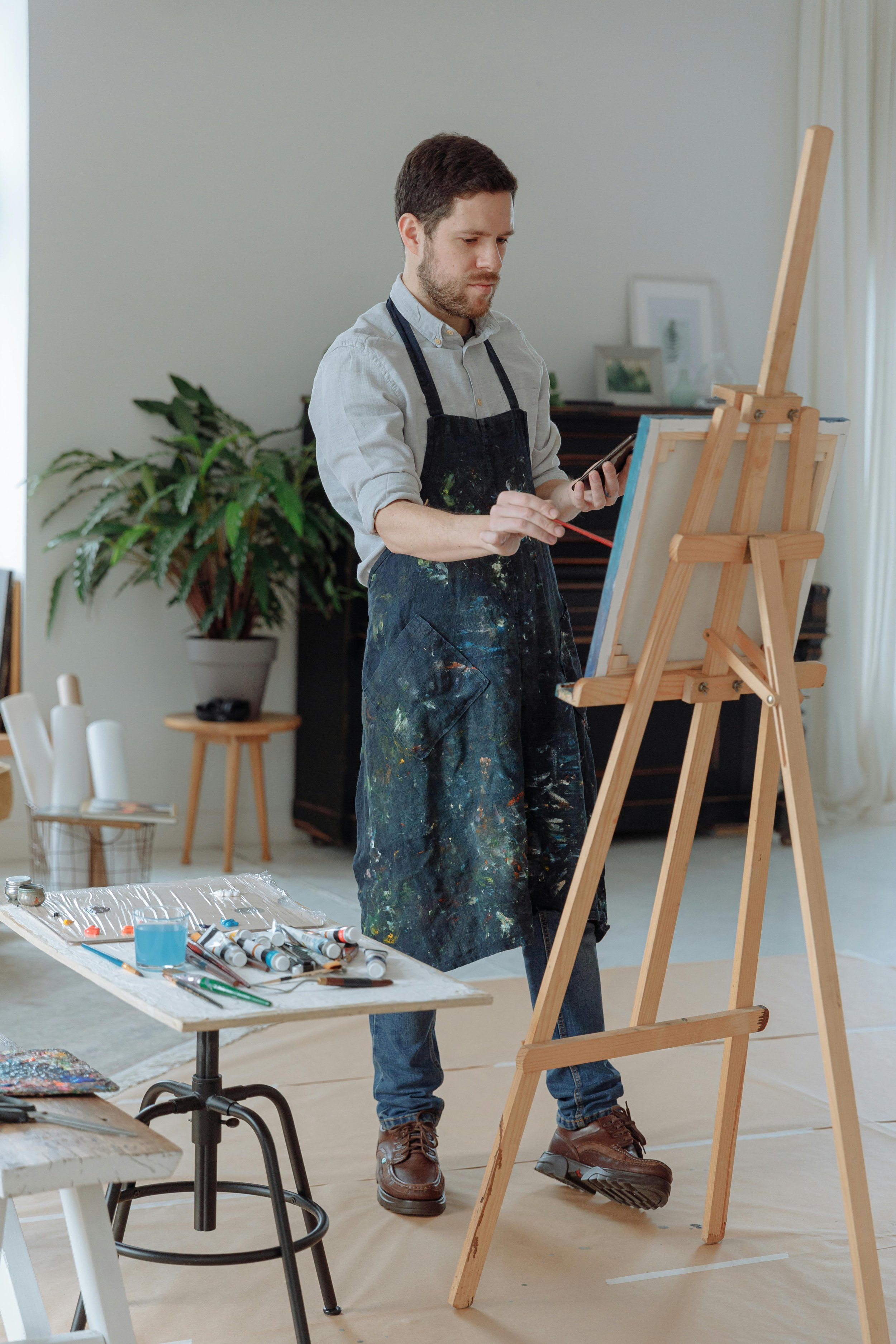 A man painting on a canvas set on an easel in a bright room, wearing a paint-splattered apron.