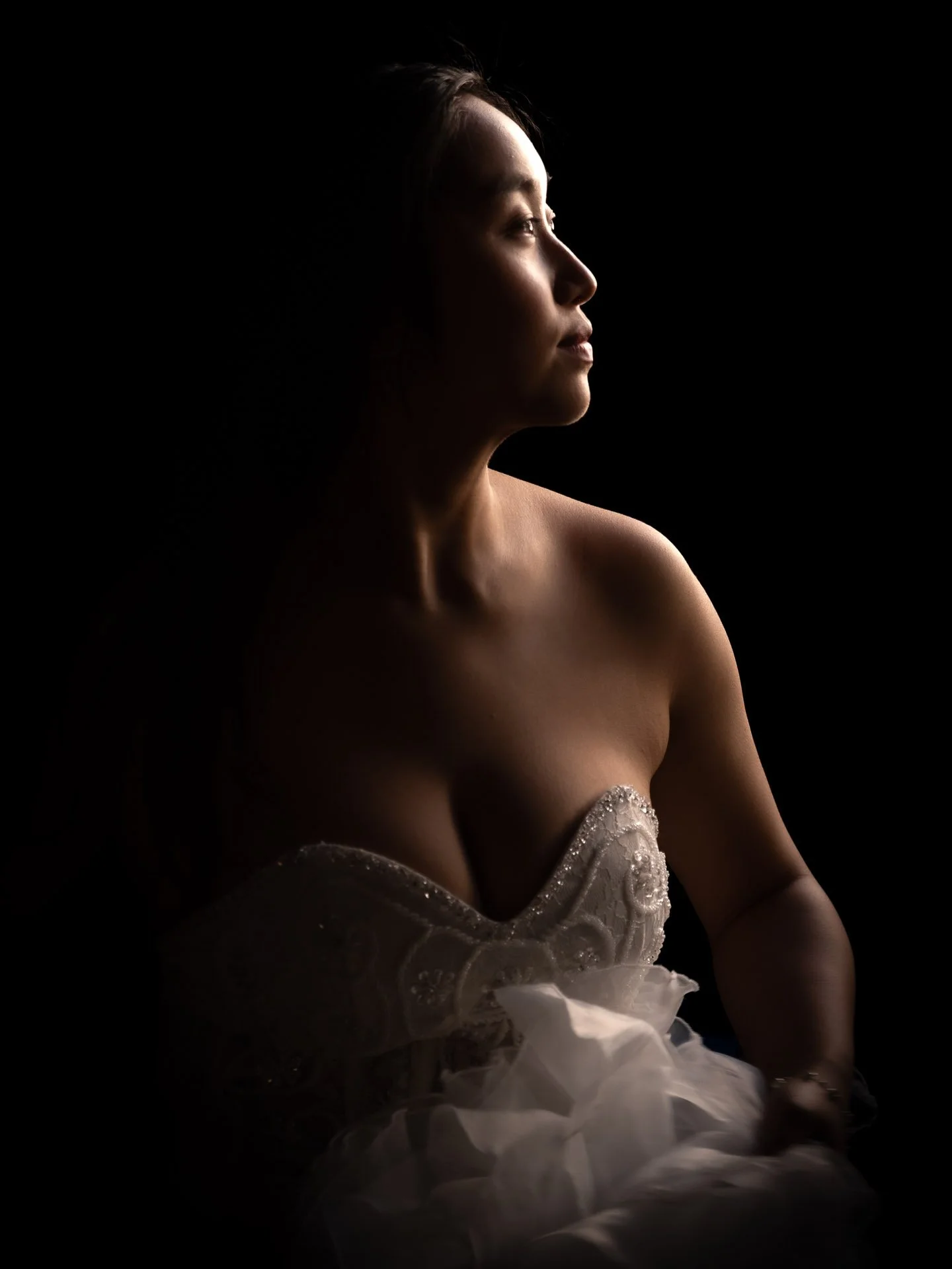 Profile of a woman with illuminated face and shoulders, wearing a strapless white dress with intricate lace details, against a black background.
