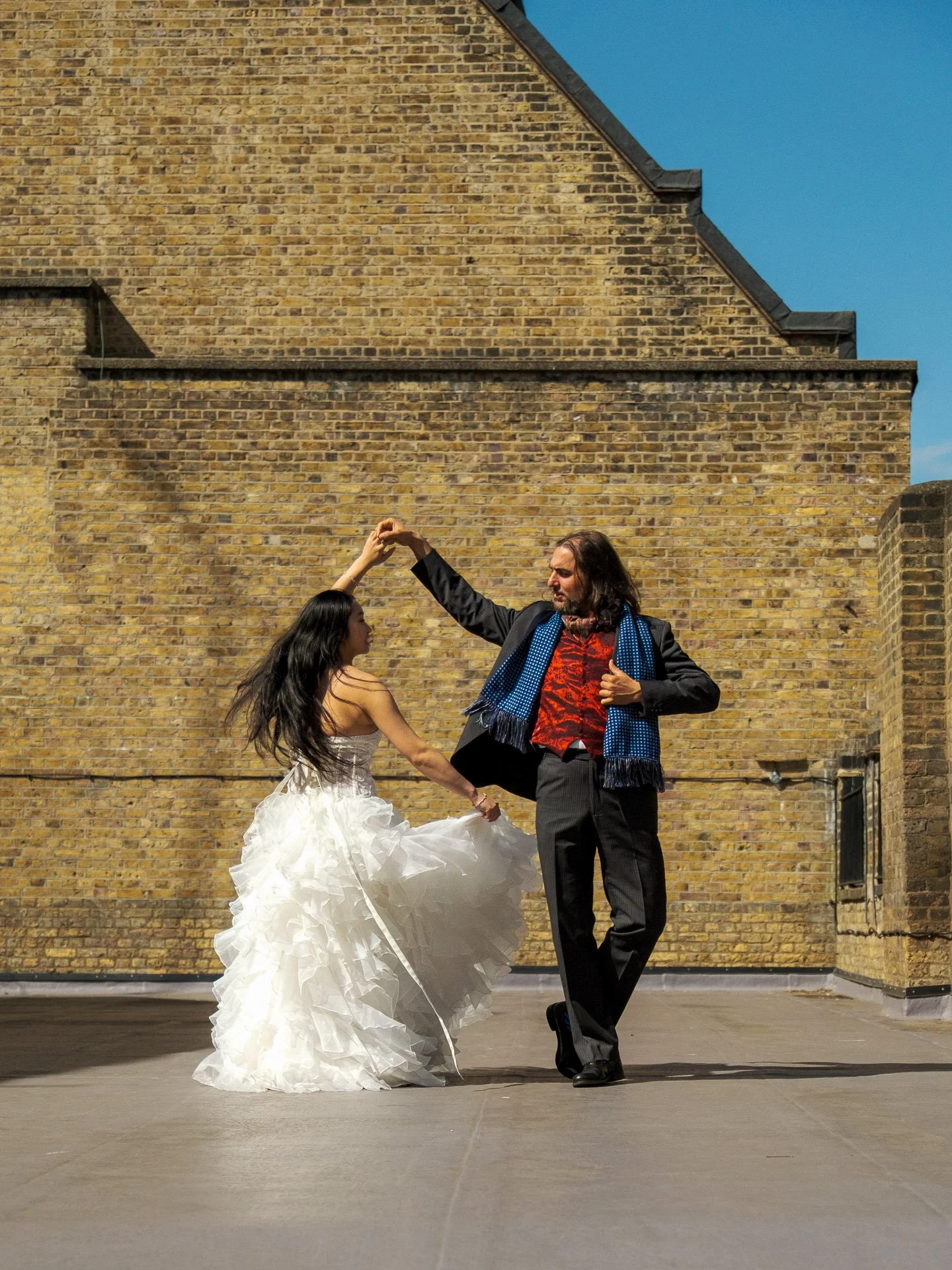 A man and woman dancing on a rooftop with a brick wall and blue sky in the background. The woman is wearing a white wedding dress, and the man is dressed in a black suit with a scarf.