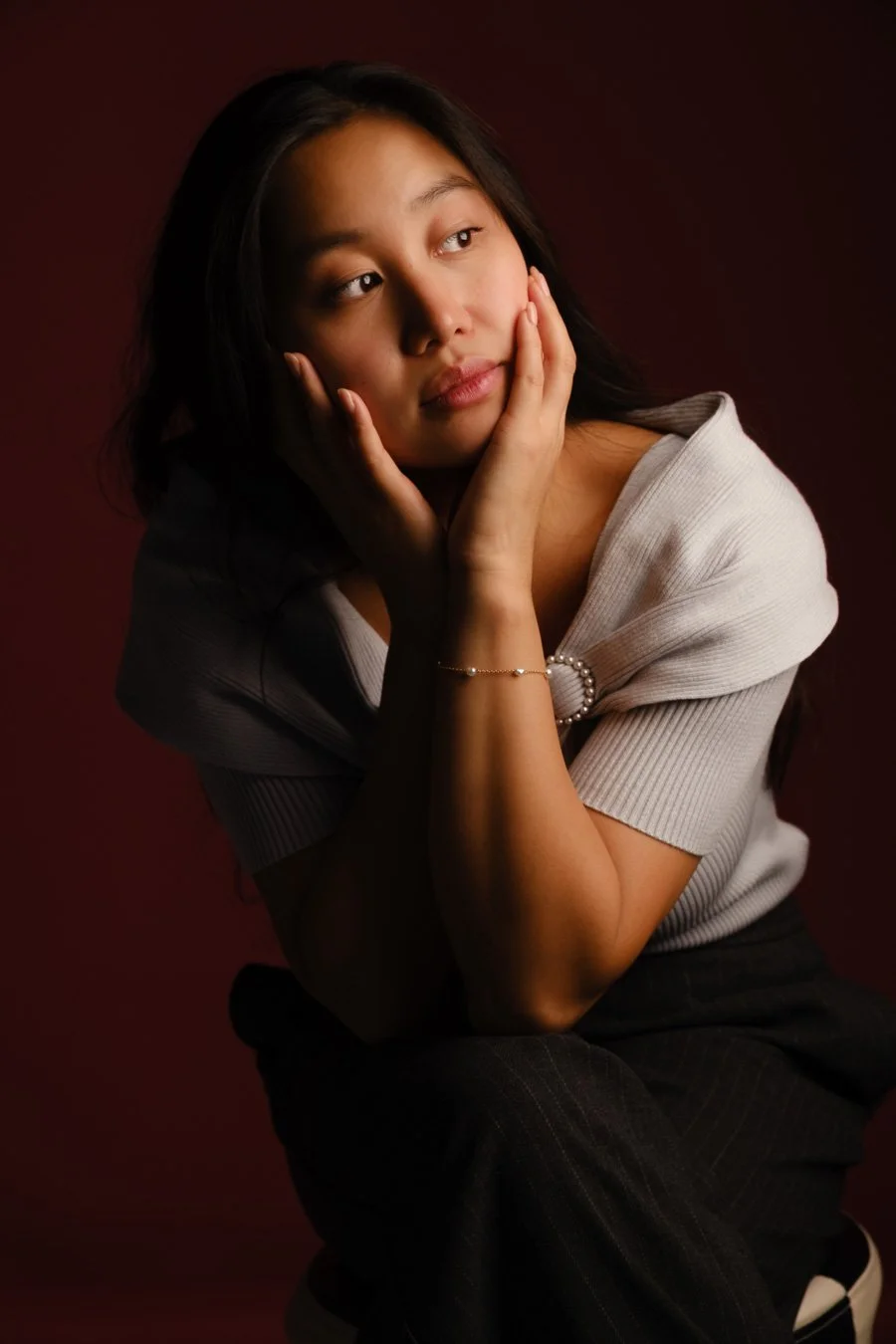 A young woman with long dark hair, wearing a light-colored top with a pearl bracelet, looks thoughtfully to the side with her chin resting in her hands against a dark background.