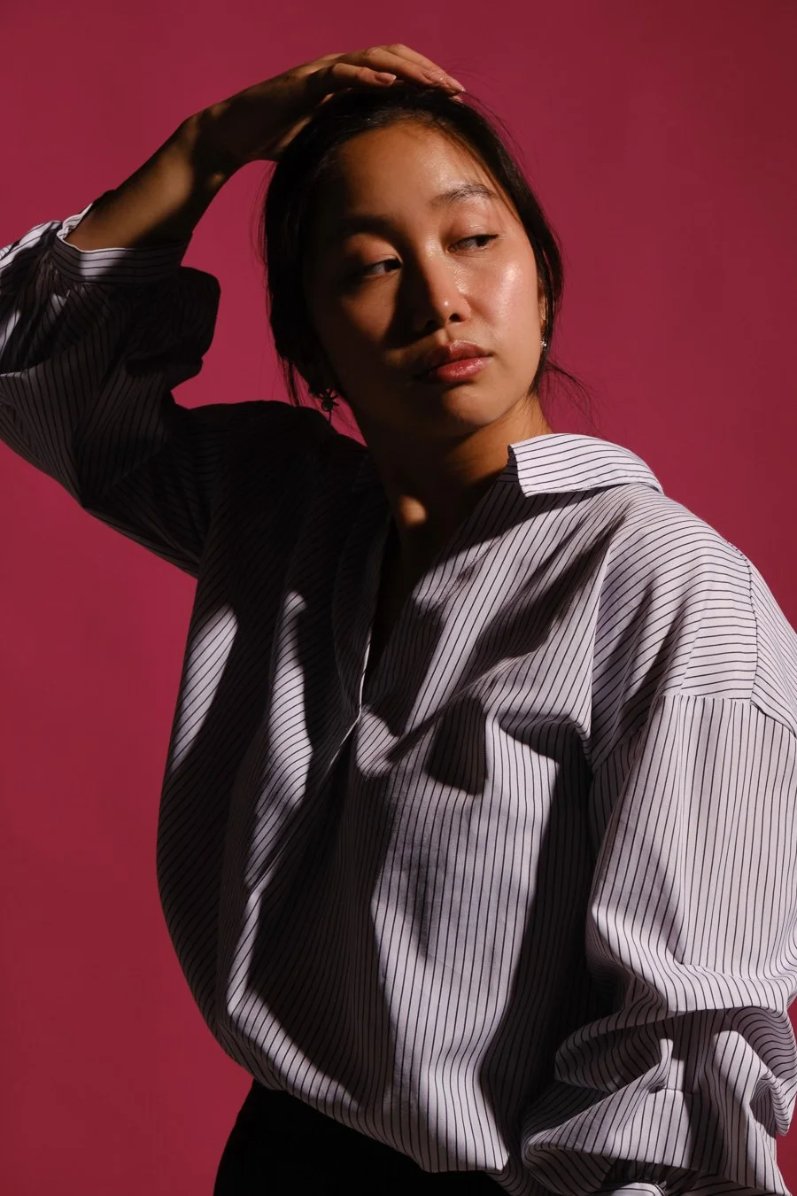 A woman with dark hair in a black and white striped shirt, posing with her hand on her head against a pink background.