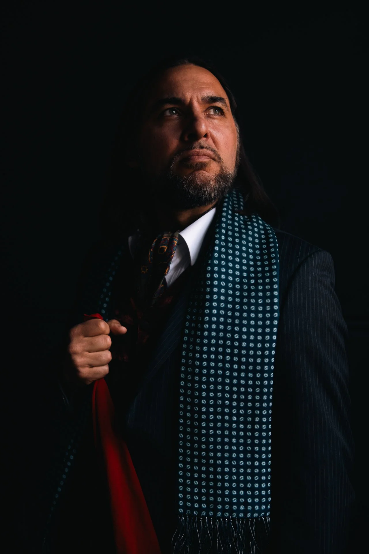 A man with long black hair, a beard, and a serious expression looking upwards, dressed in a dark pinstripe suit with a white shirt, colorful tie, and a patterned blue scarf, holding the lapel of his suit against a dark background.