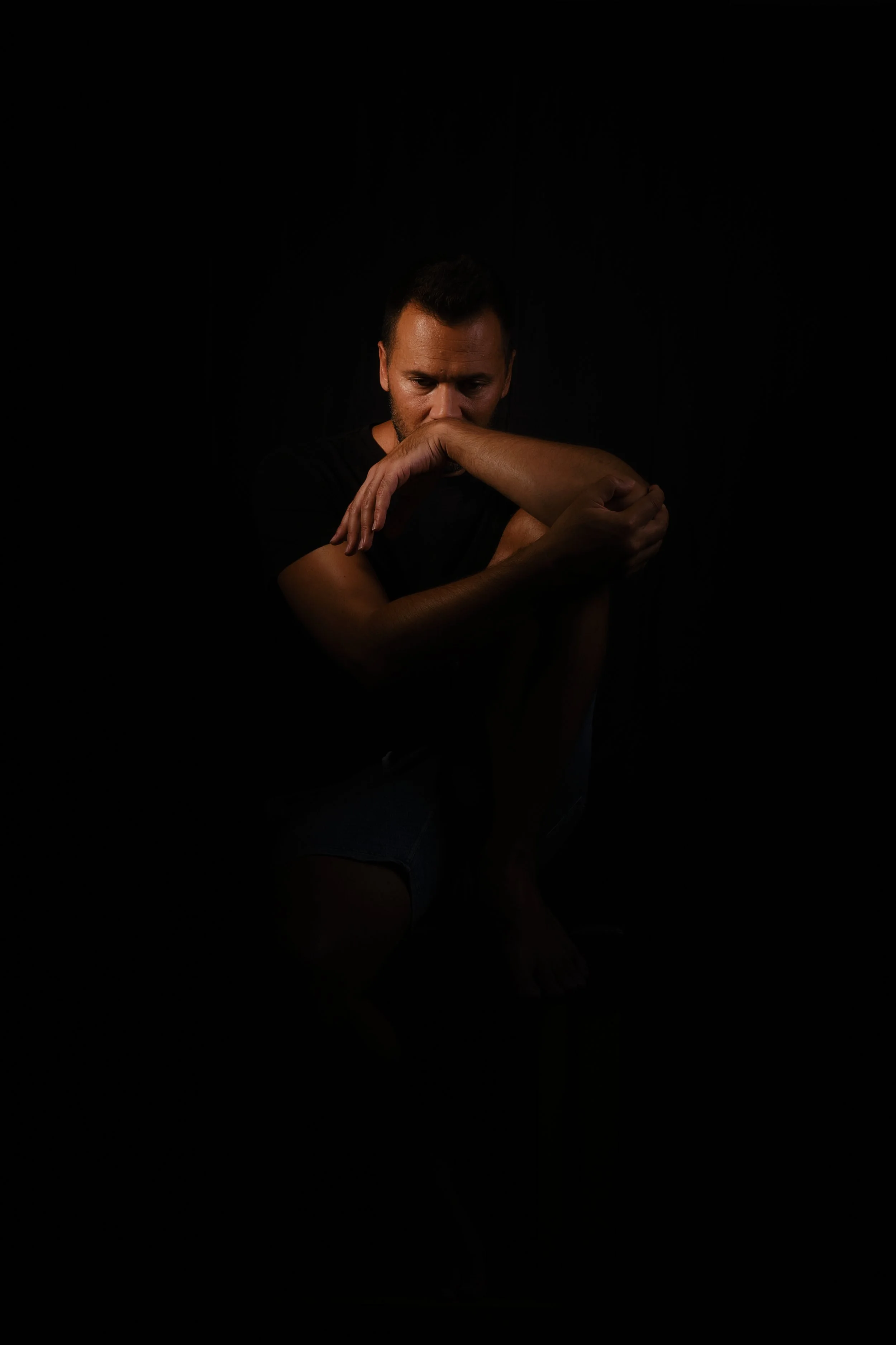 A man sitting in darkness, hugging his knees with his right arm, looking down thoughtfully.