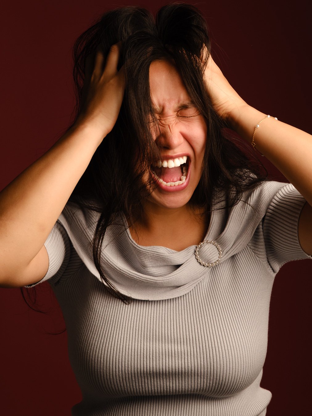 A woman with dark hair holding her head and screaming or yelling, appears distressed or overwhelmed against a dark red background.