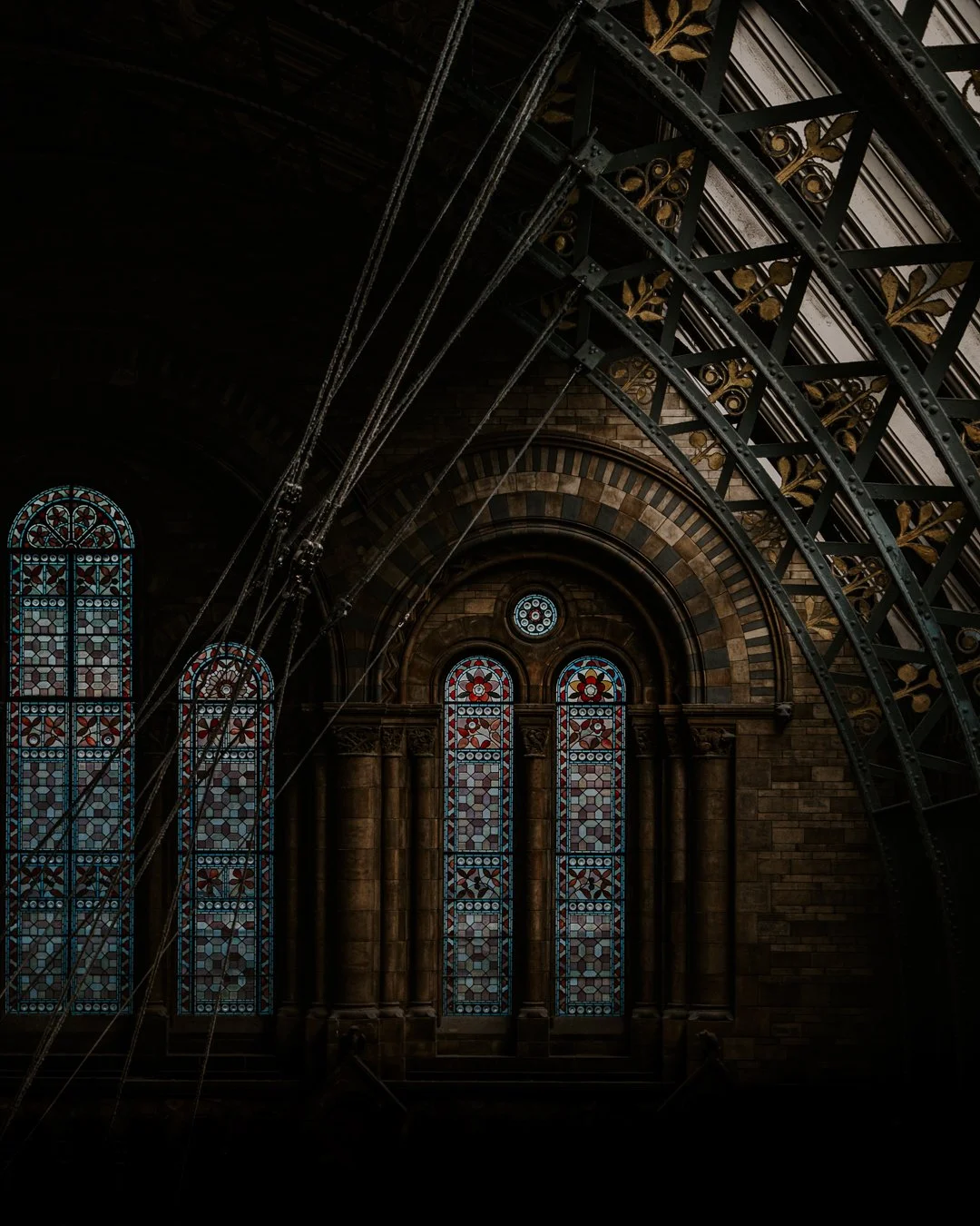 Inside a dimly lit church with three stained glass windows with geometric and floral patterns, pointed arch frames, brick walls, and decorative metalwork overhead.