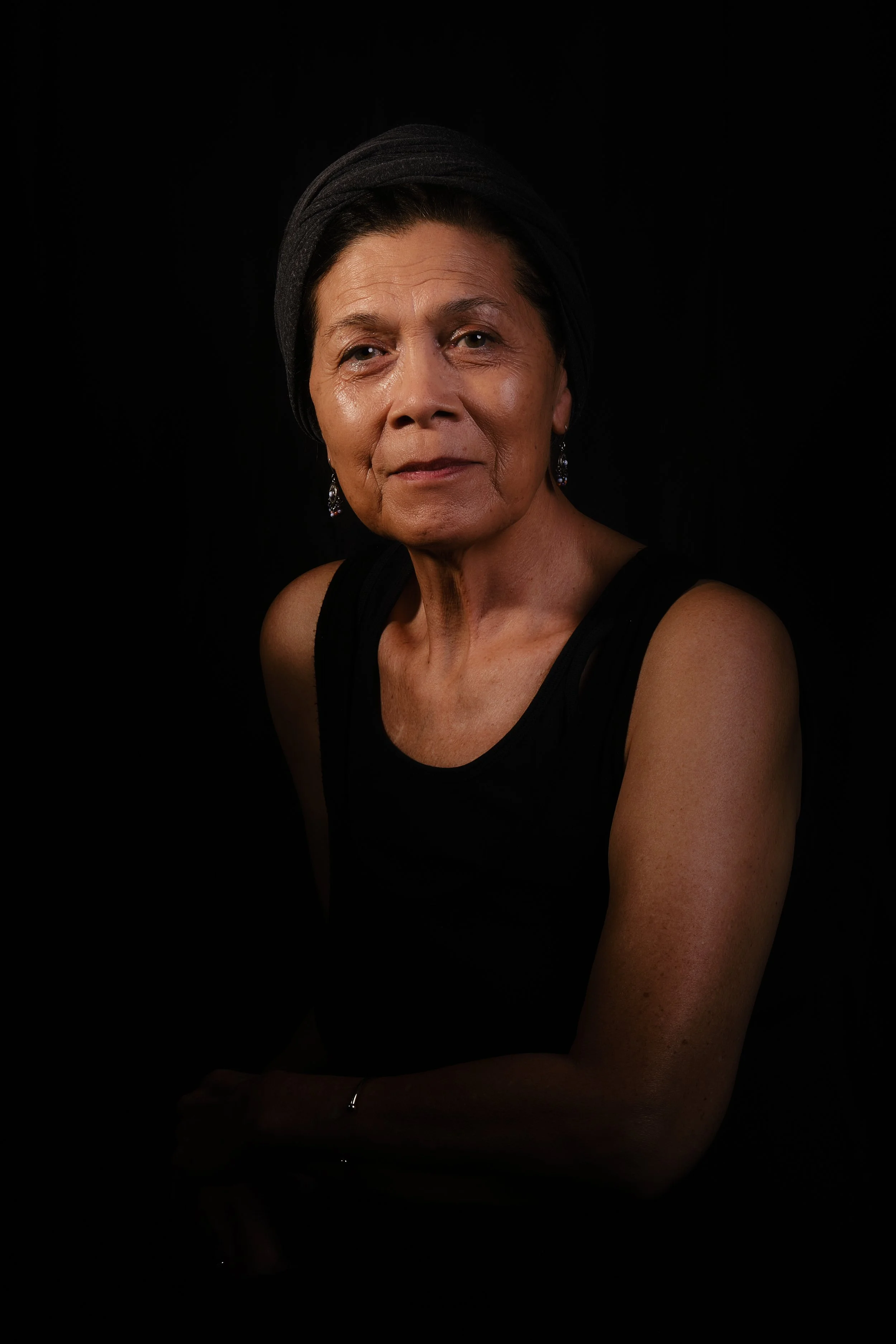 An elderly woman with short dark hair, wearing a black headscarf and black sleeveless top, sitting against a dark background.