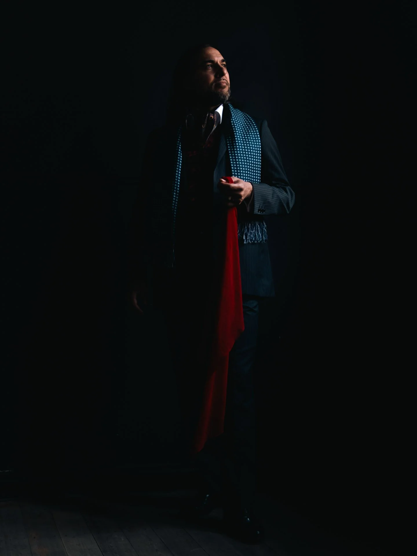 A man in a dark suit holding a red object, facing slightly to the right against a dark background.