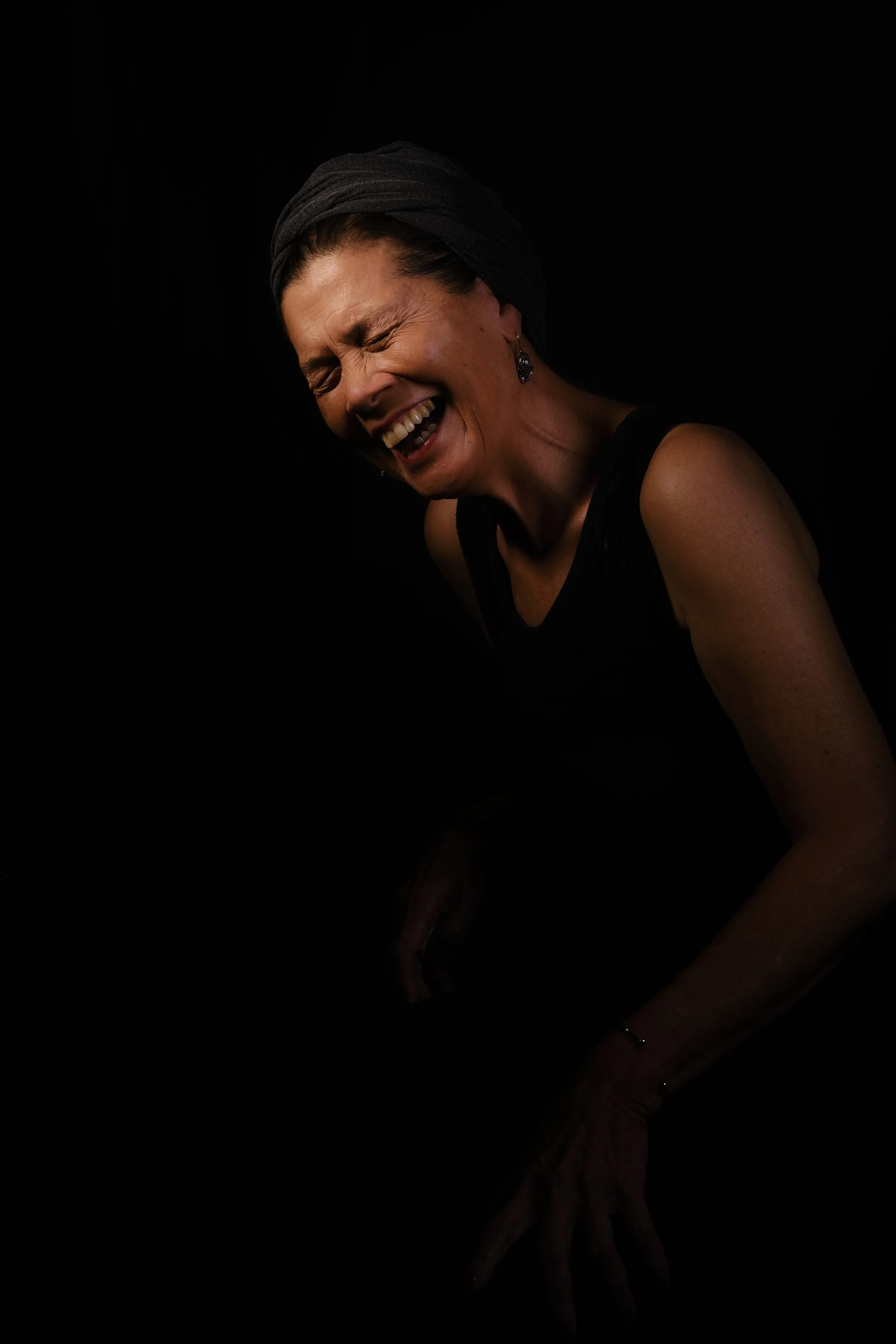 A woman wearing a dark headscarf and sleeveless top is laughing with her eyes closed, against a black background.