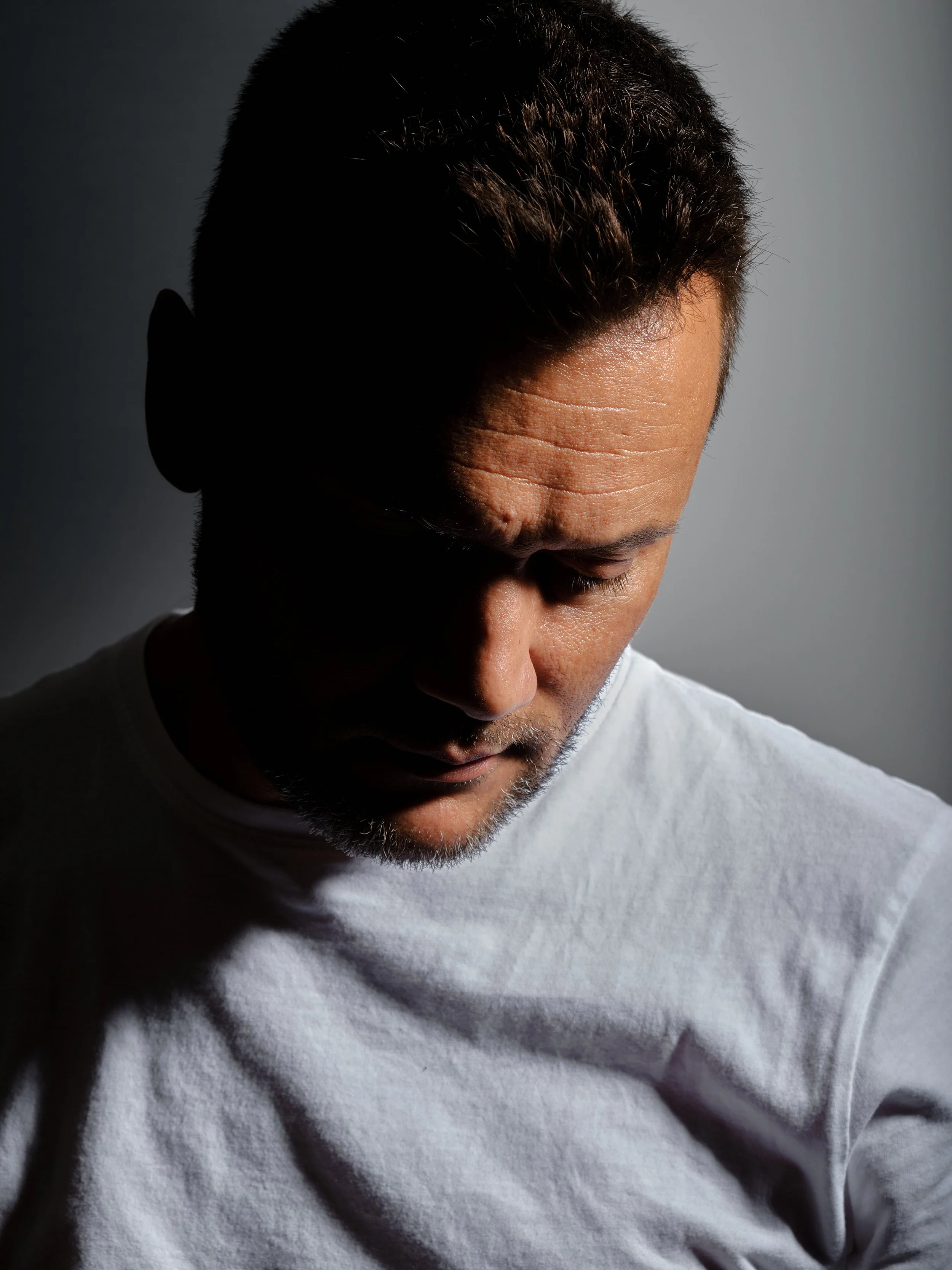 A man with short dark hair, slightly greying, wearing a white t-shirt, looking down with a shadowed face, against a dark background