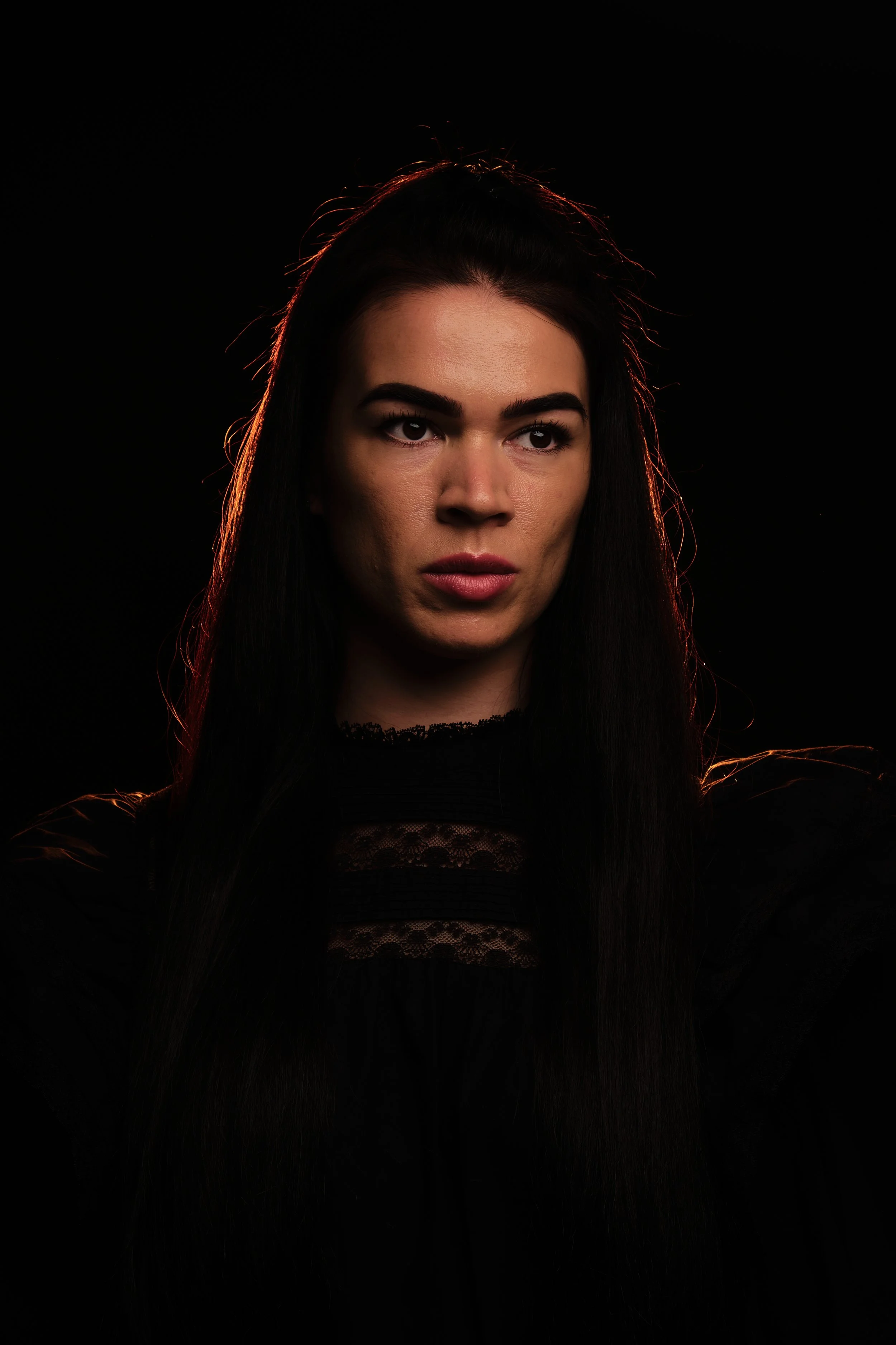 Close-up portrait of a young woman with long dark hair and bold eyebrows, wearing a black lace top, against a dark background.