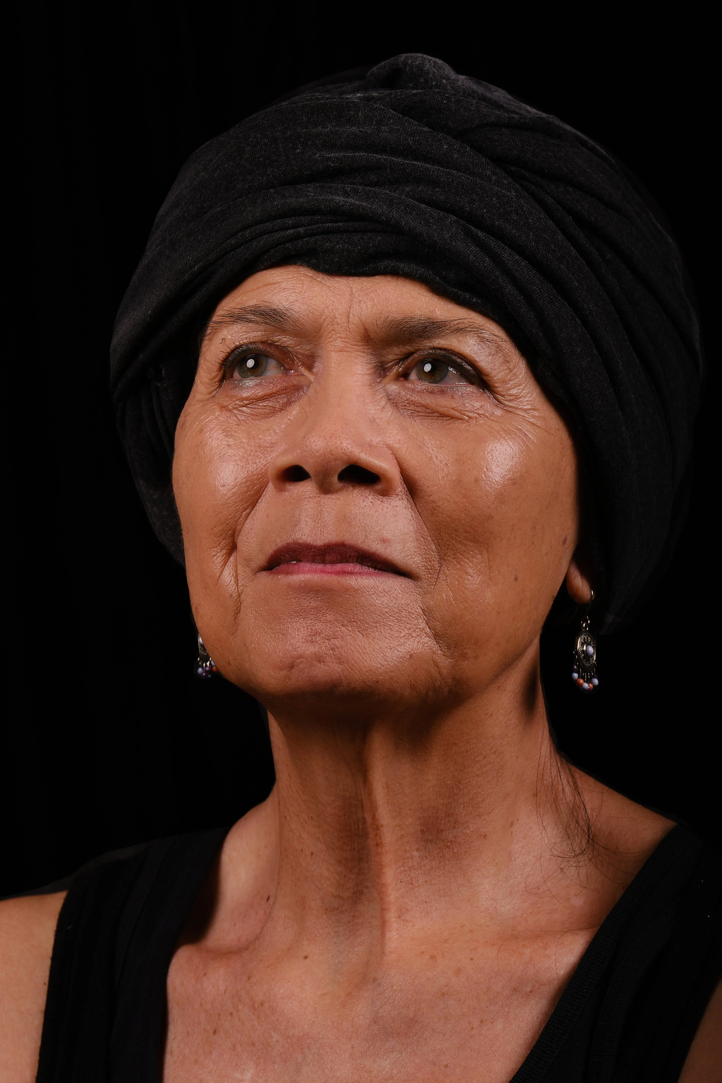 Close-up of an elderly woman with tan skin, wearing a black headwrap and earrings, against a black background.