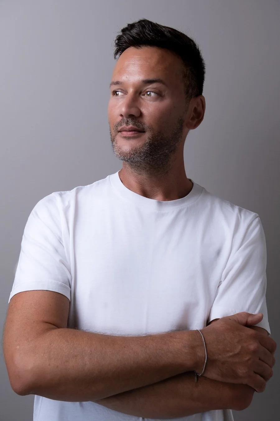 A middle-aged man with short dark hair, facial hair, wearing a plain white t-shirt, standing with arms crossed against a gray background, looking to the side.