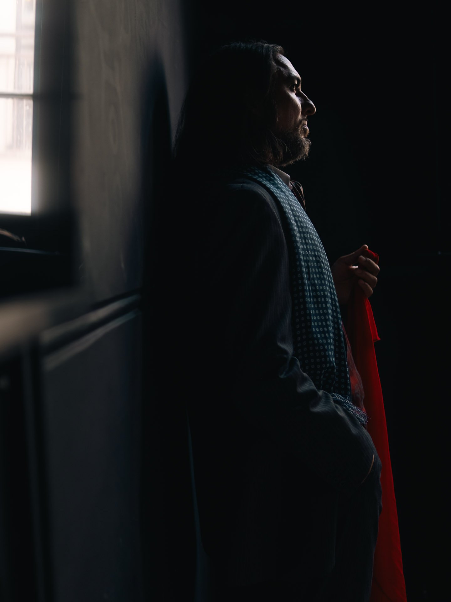 Side profile of a man with long hair and beard, standing against a dark background, holding a red cloth in his right hand, with light coming from a window