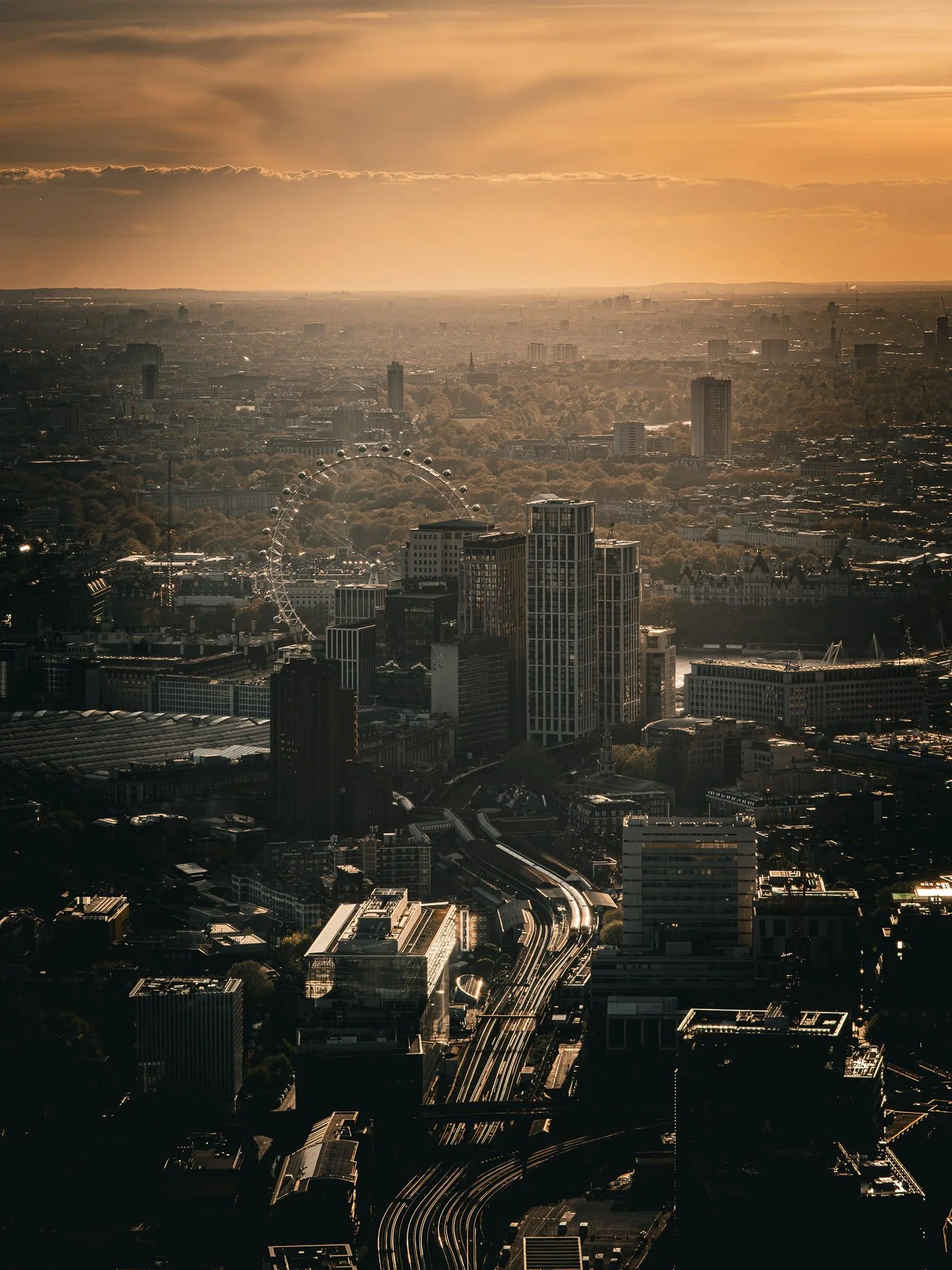 Aerial view of a cityscape at sunset with tall buildings, a Ferris wheel, and a network of train tracks.
