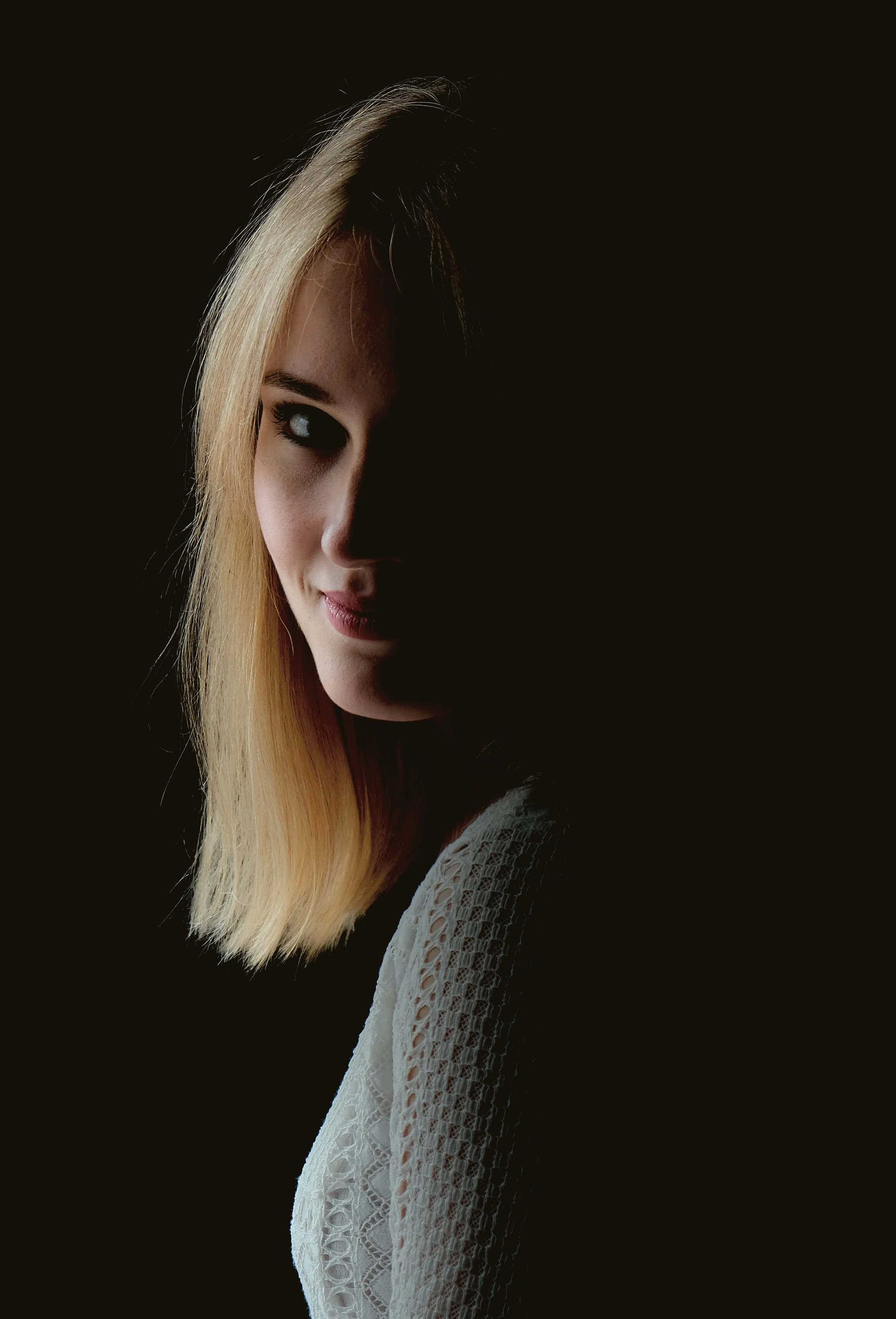 A young woman with blonde hair partly shadowed, wearing a white lacy top, looking at the camera with a slight smile against a dark background.