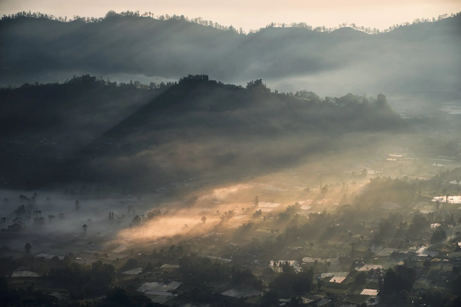 Aerial view of a mountain landscape with fog and sunlight illuminating the valley below, showing hills and scattered farms or greenhouses.