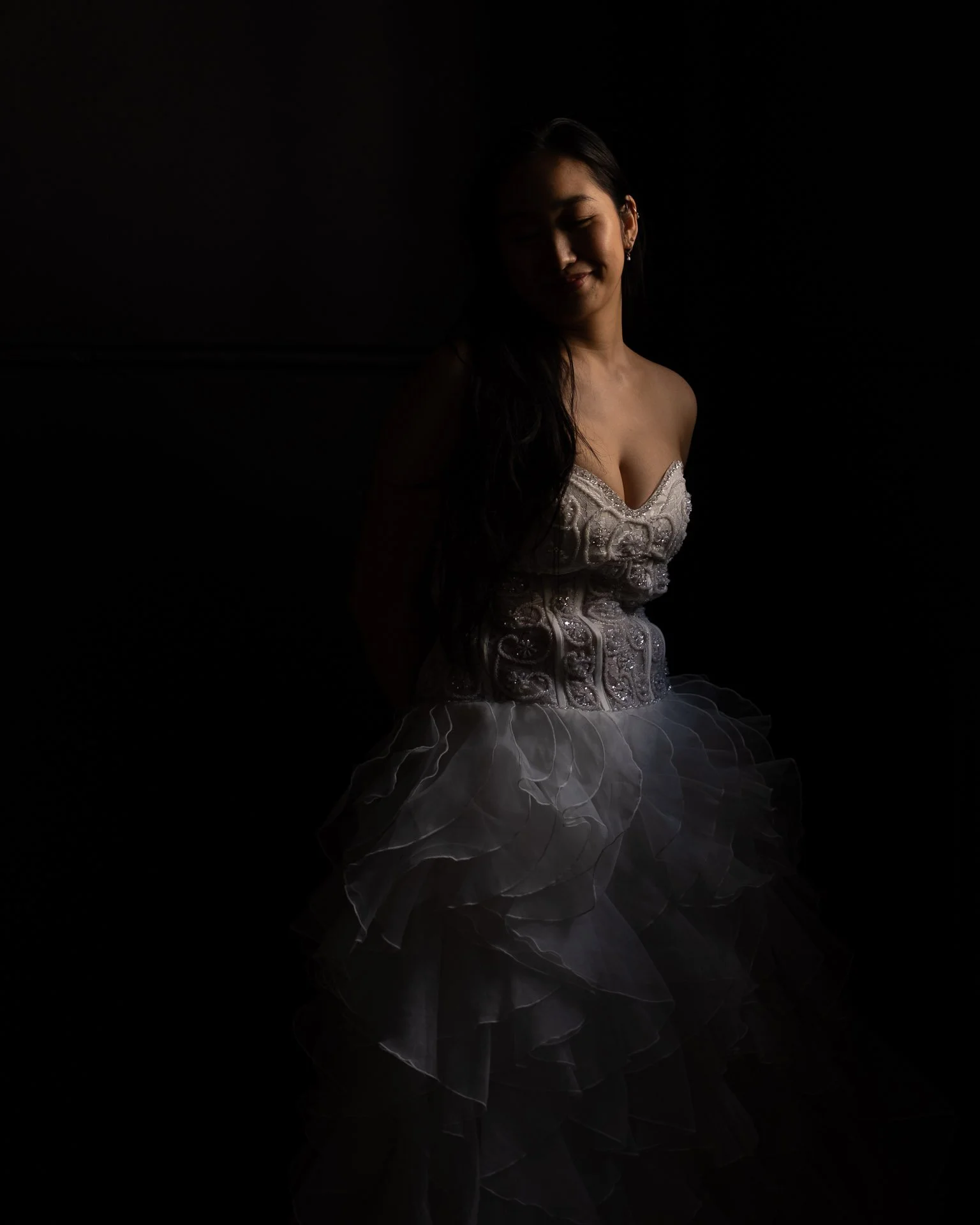 A woman in an intricate white embroidered dress with ruffled layers, standing in a dark setting with soft lighting highlighting her face and dress.