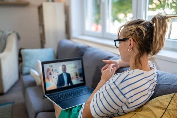 Woman sitting on a couch, wearing glasses and a striped shirt, participating in a video call on her laptop in a cozy living room.