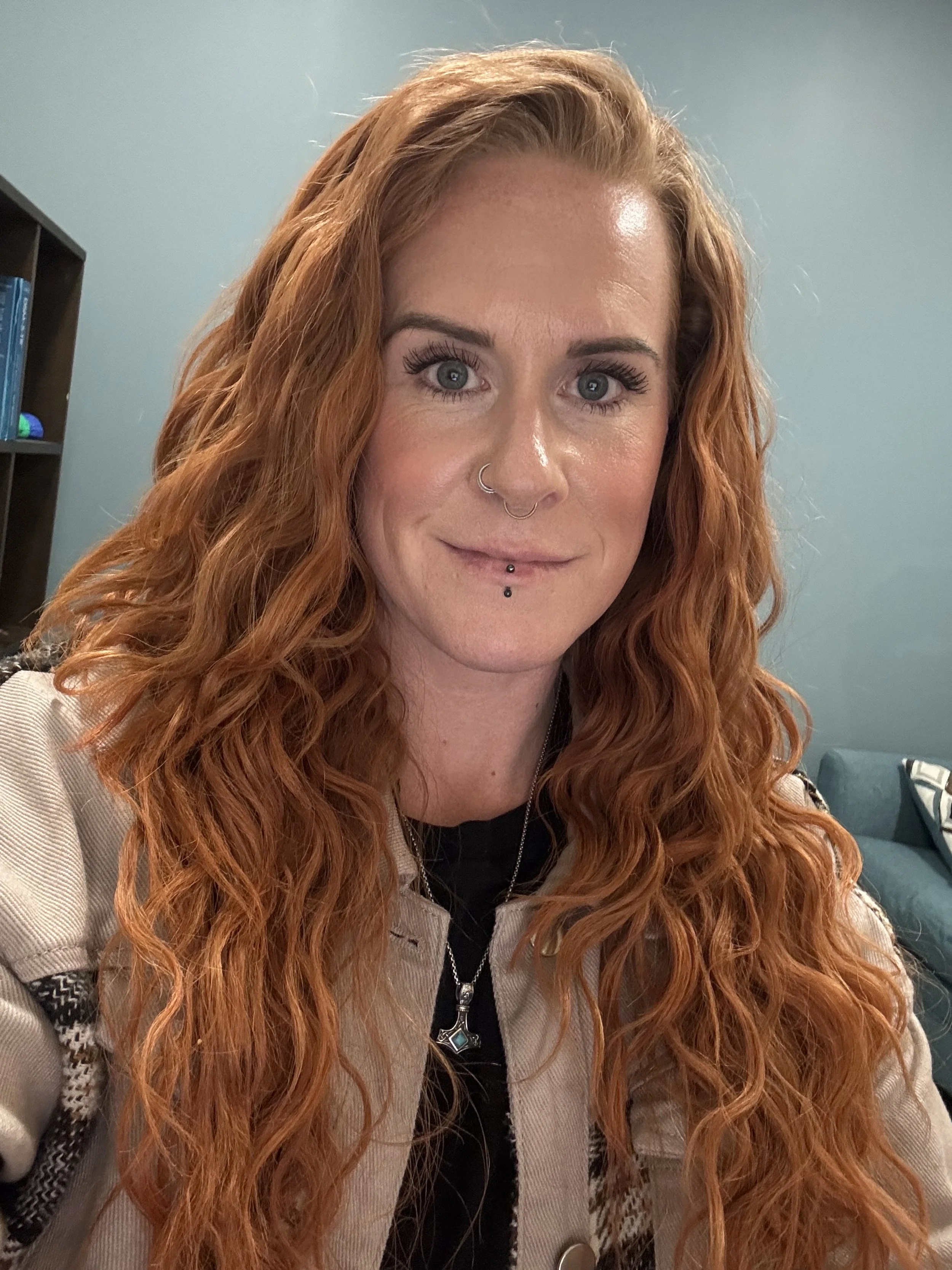 A woman with long curly red hair, light skin, blue eyes, and multiple facial piercings, wearing a beige jacket and a necklace, taking a selfie in a room with a blue wall and bookshelf.