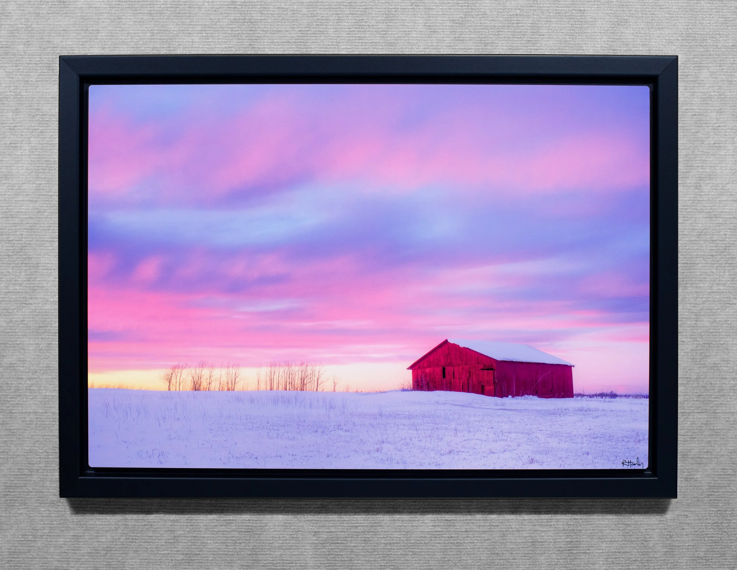 A framed landscape photo of a snow-covered field with a red barn and bare trees in the distance against a pink and purple sky at sunset or sunrise.