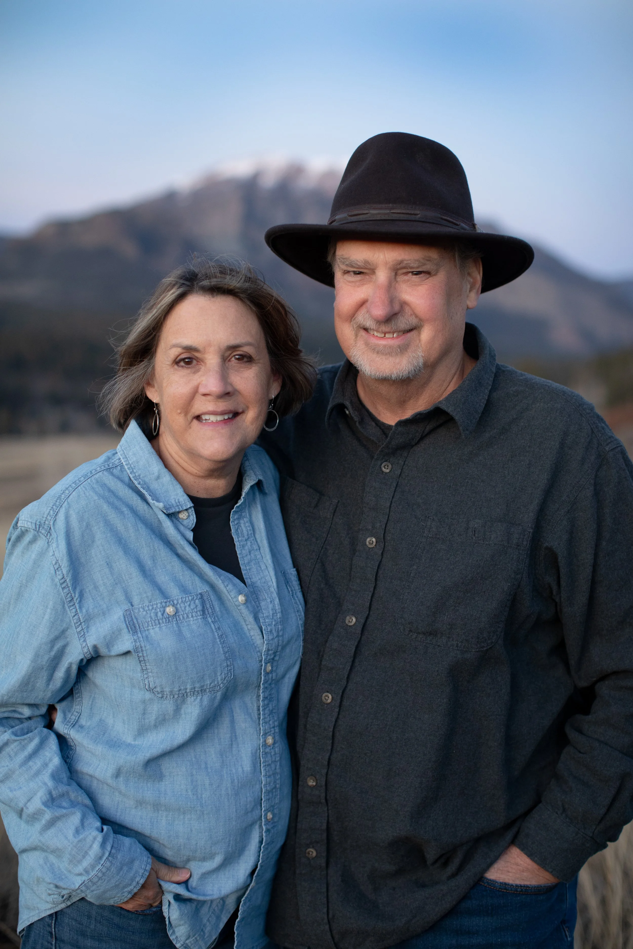 A middle-aged couple standing outdoors, smiling, with mountains in the background. The woman is wearing a denim shirt and has short brown hair, and the man is wearing a black hat and a dark shirt.