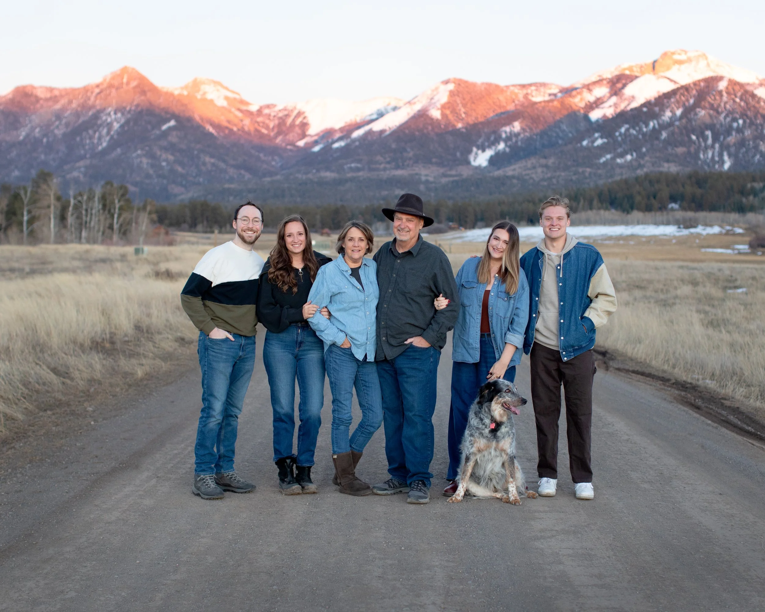 A group of six people and a dog standing on a dirt road with mountains and a grassy field in the background.
