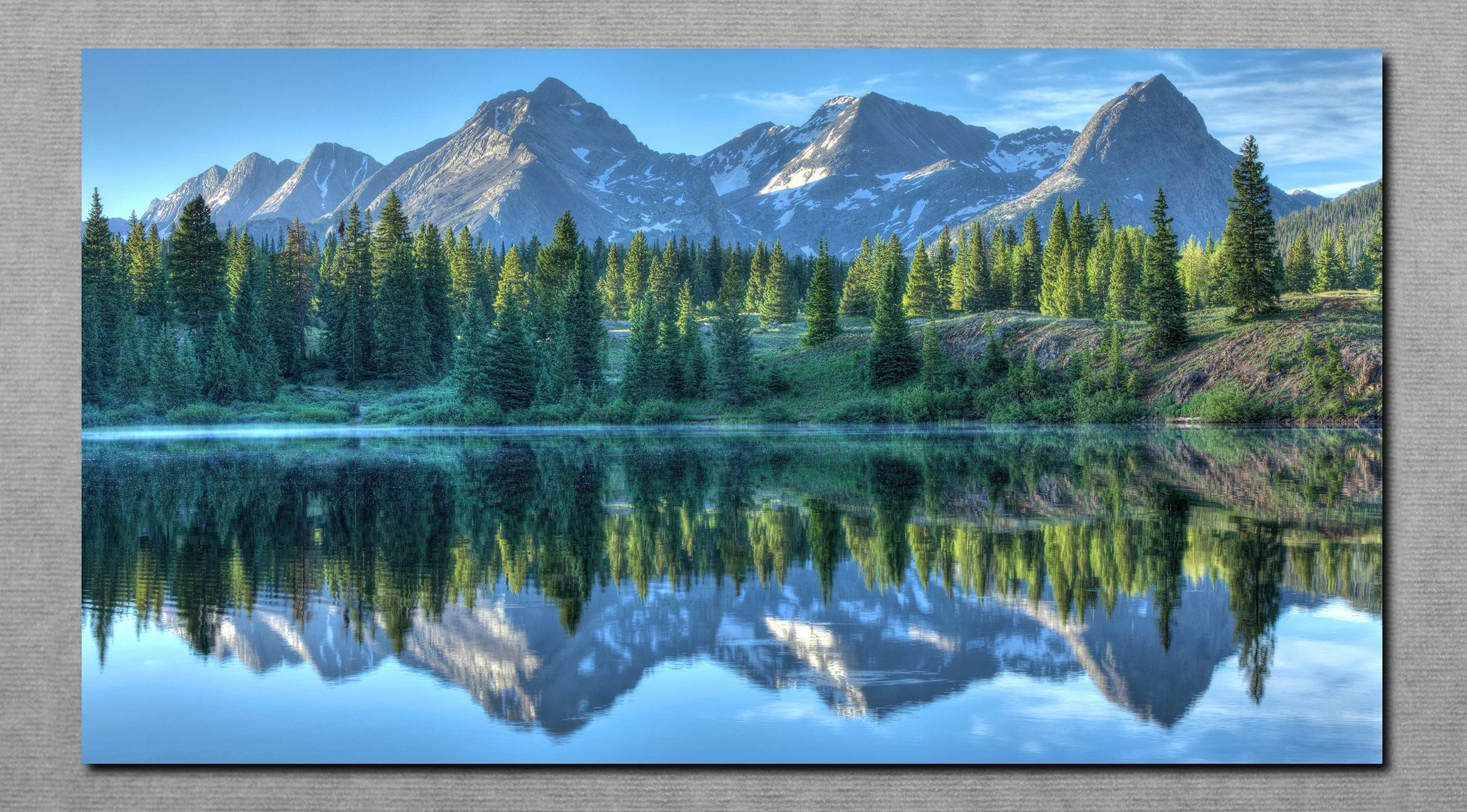Scenic view of snow-capped mountains reflected in a calm lake surrounded by pine trees under a blue sky.