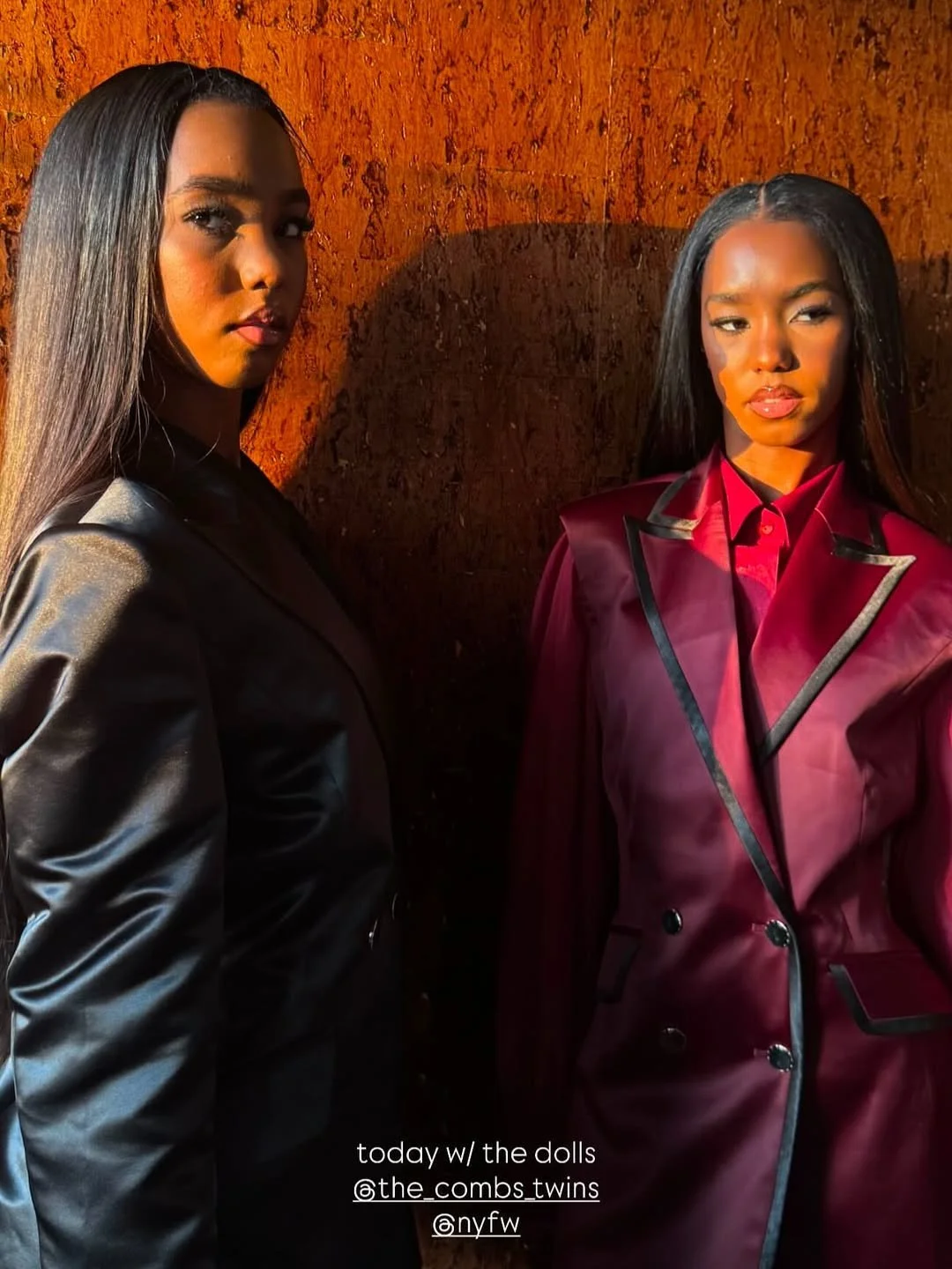 Two women with straight black hair standing against a textured orange wall, dressed in shiny leather jackets, one black and one deep red.