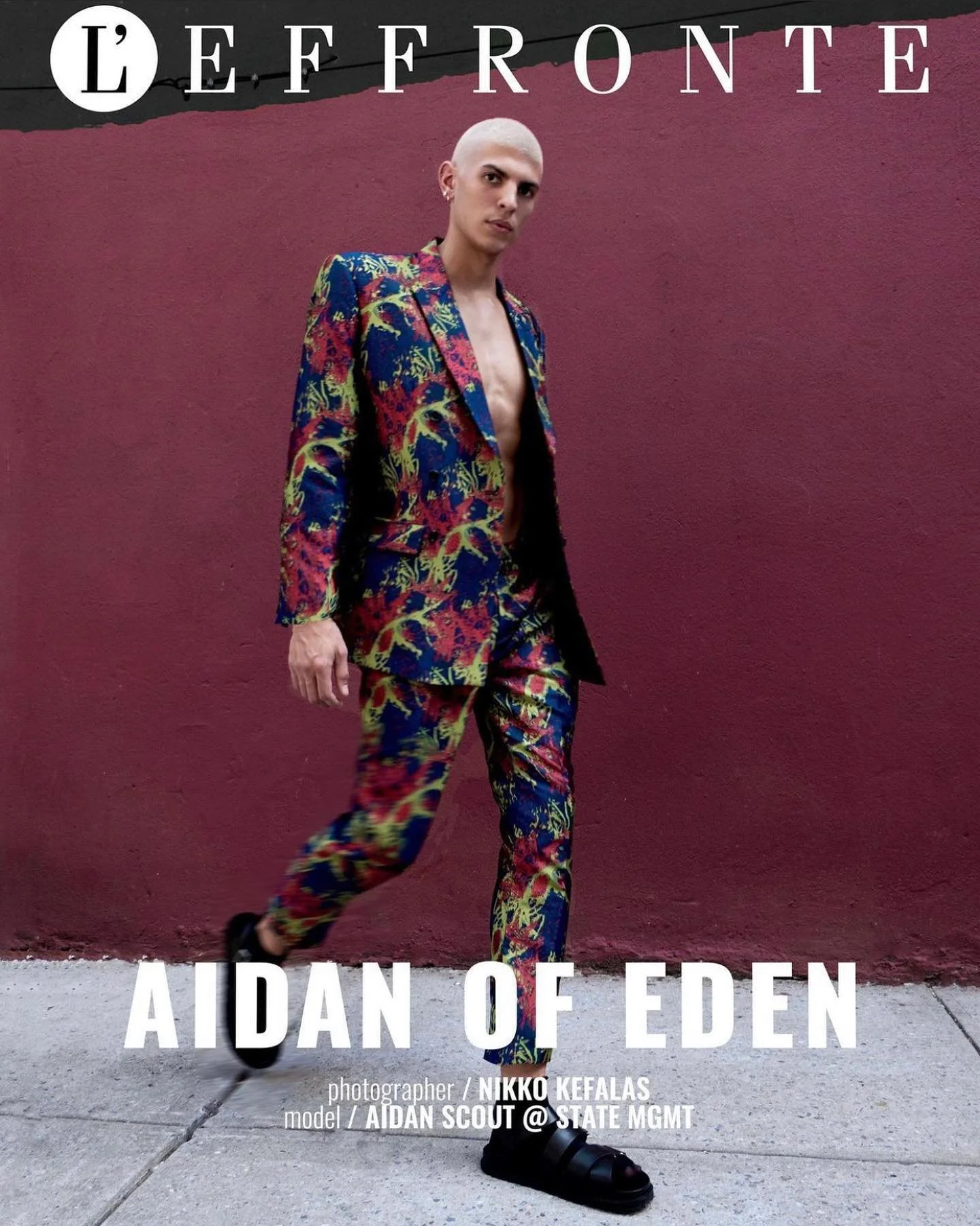 A person with a shaved head wearing a colorful, patterned suit walks in front of a dark pink wall.