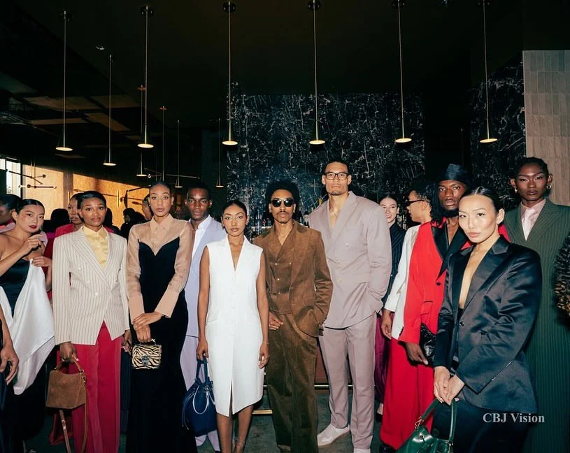 Group of diverse stylish people at a fashion event or gallery, standing in a line against a dark, marble-textured wall, with some holding handbags.
