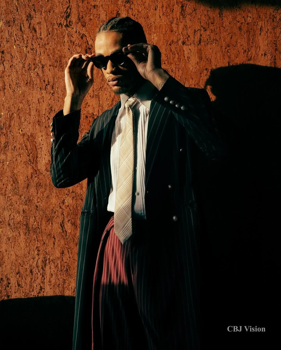 A man with braided hair adjusting sunglasses against a textured orange background, wearing a pinstripe suit, a light-colored tie, and a white shirt.