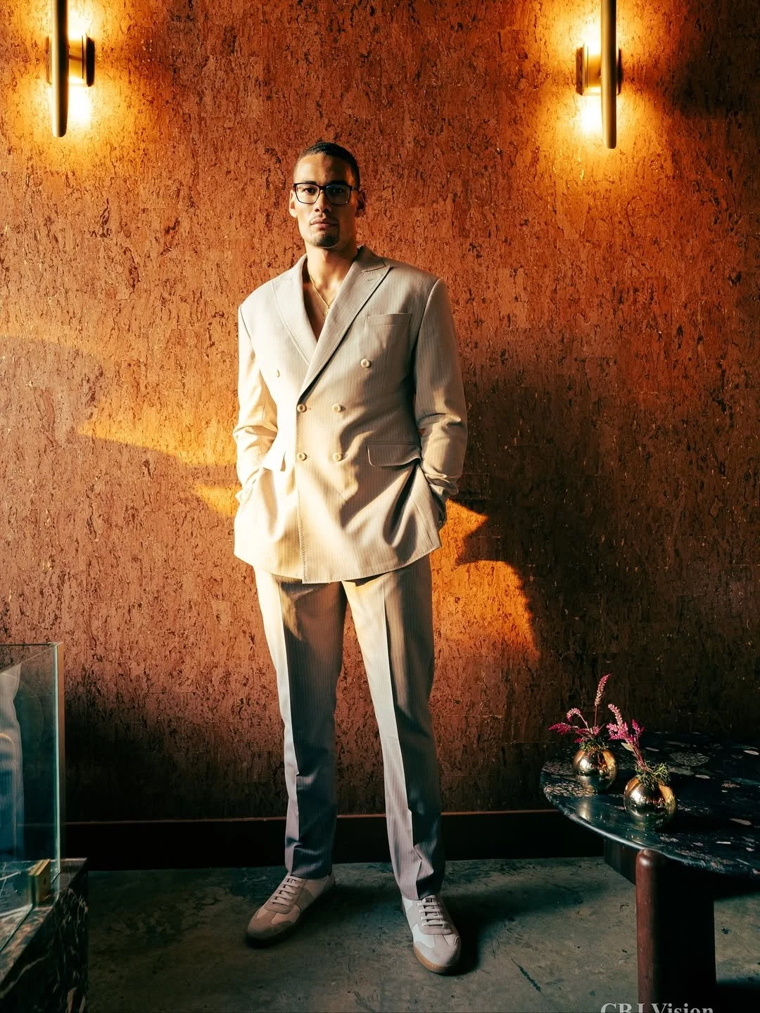 Man in a cream-colored double-breasted suit with a pocket on the left chest, standing with hands in pockets against a textured orange wall illuminated by wall-mounted lights, with a small dark table holding two vases with pink flowers to his right.