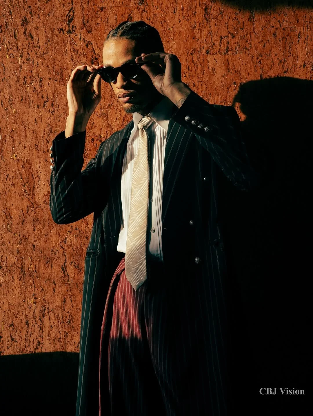 A man wearing a pinstripe suit, light-colored shirt, and tie, adjusting sunglasses against a textured red wall, with a shadow cast on the wall.