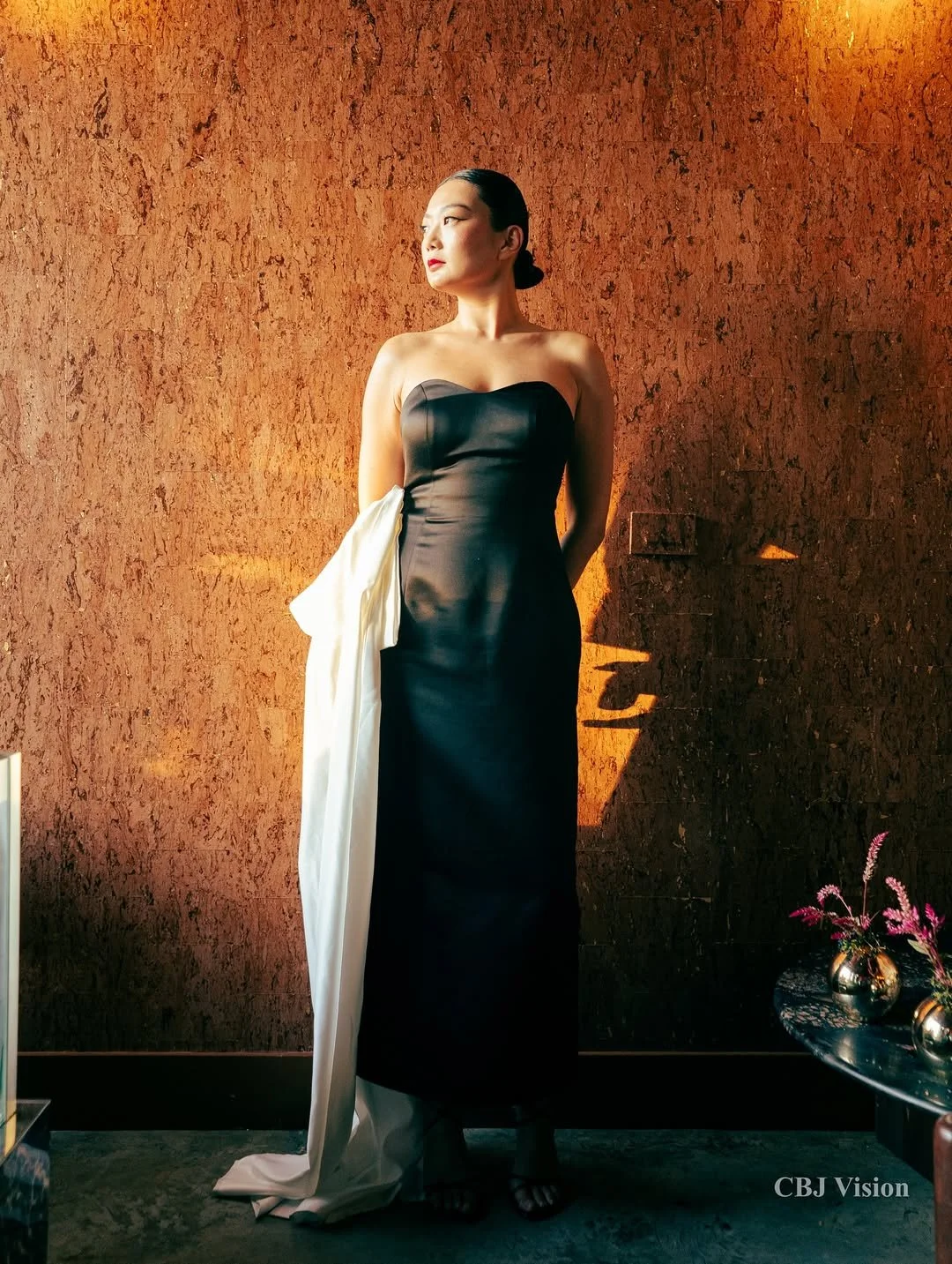 A woman in a black strapless gown with a white shawl draped over one arm standing against a textured brown wall, illuminated by warm lighting.