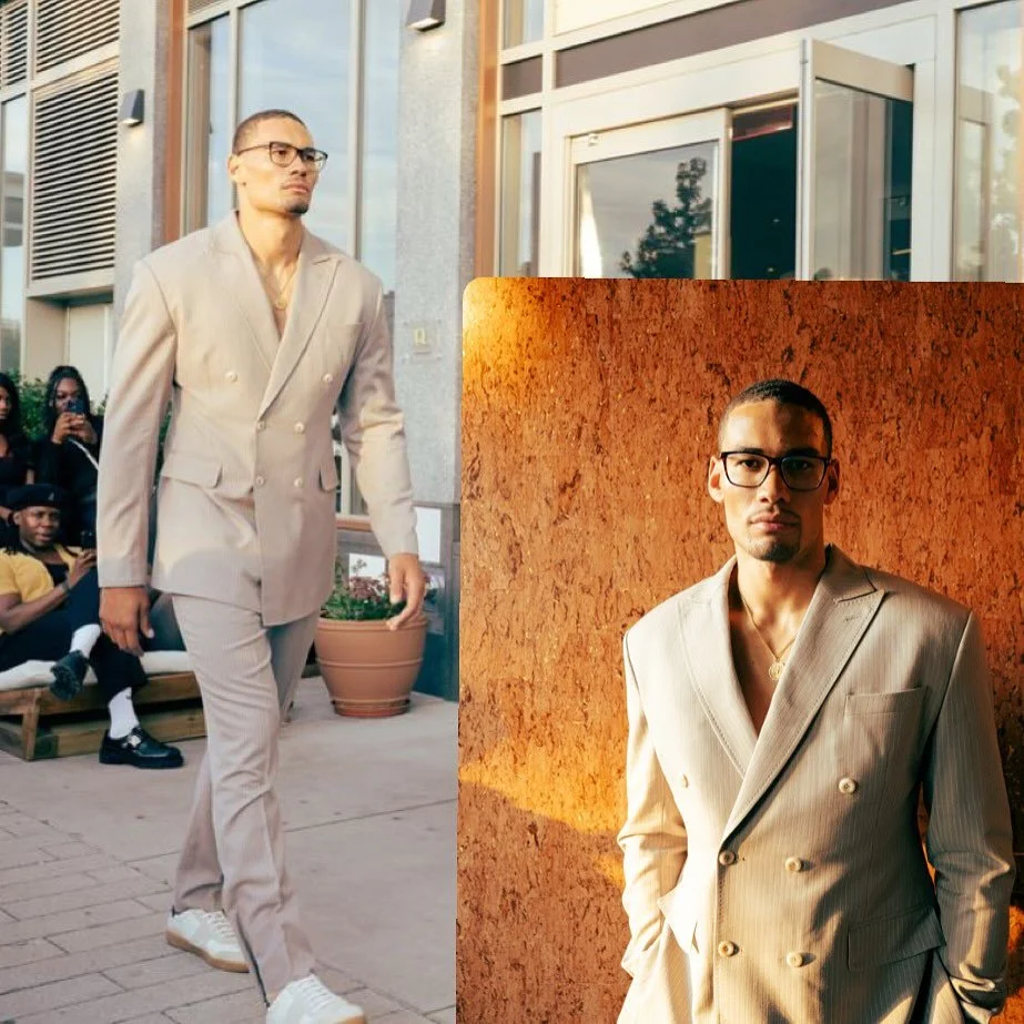 A man with glasses and a beige double-breasted suit with wide lapels, standing outdoors in front of a building. In the background, there are people sitting on benches, and large glass windows and door of the building.