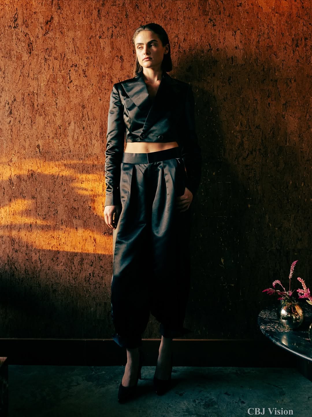 A woman standing against a textured brown wall, wearing a black satin crop top and matching high-waisted pants, with her left hand in her pocket and looking to the side.