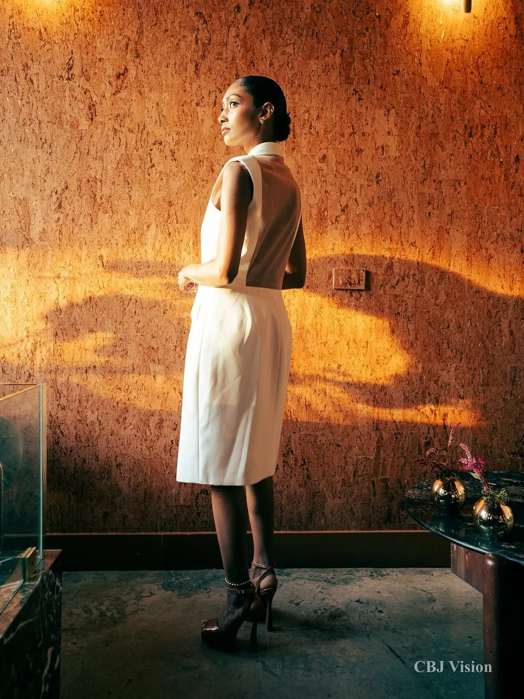 A woman in elegant beige clothing and high heels standing near a textured orange wall with spotlighting.