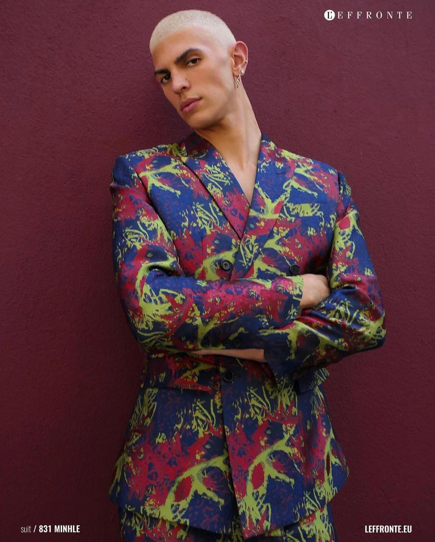 Person with short platinum blonde hair wearing a colorful abstract patterned suit with arms crossed, standing against a dark pink wall.
