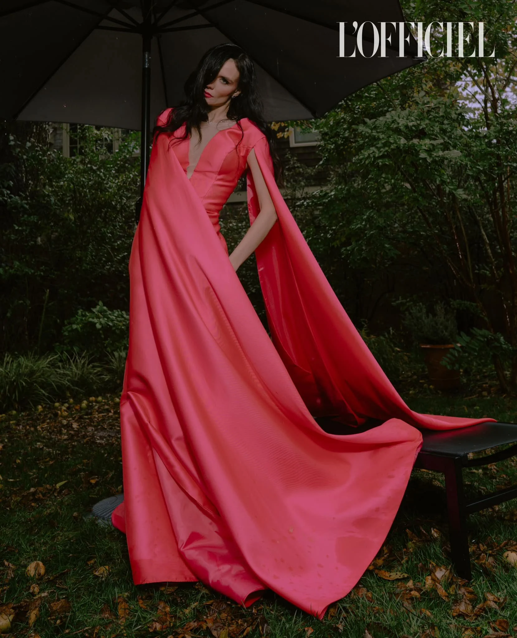 A woman in a long, flowing red dress standing outdoors under a black umbrella. She has dark hair and is standing in a garden with green foliage and a wooden house in the background. The image is from L'Officiel magazine.