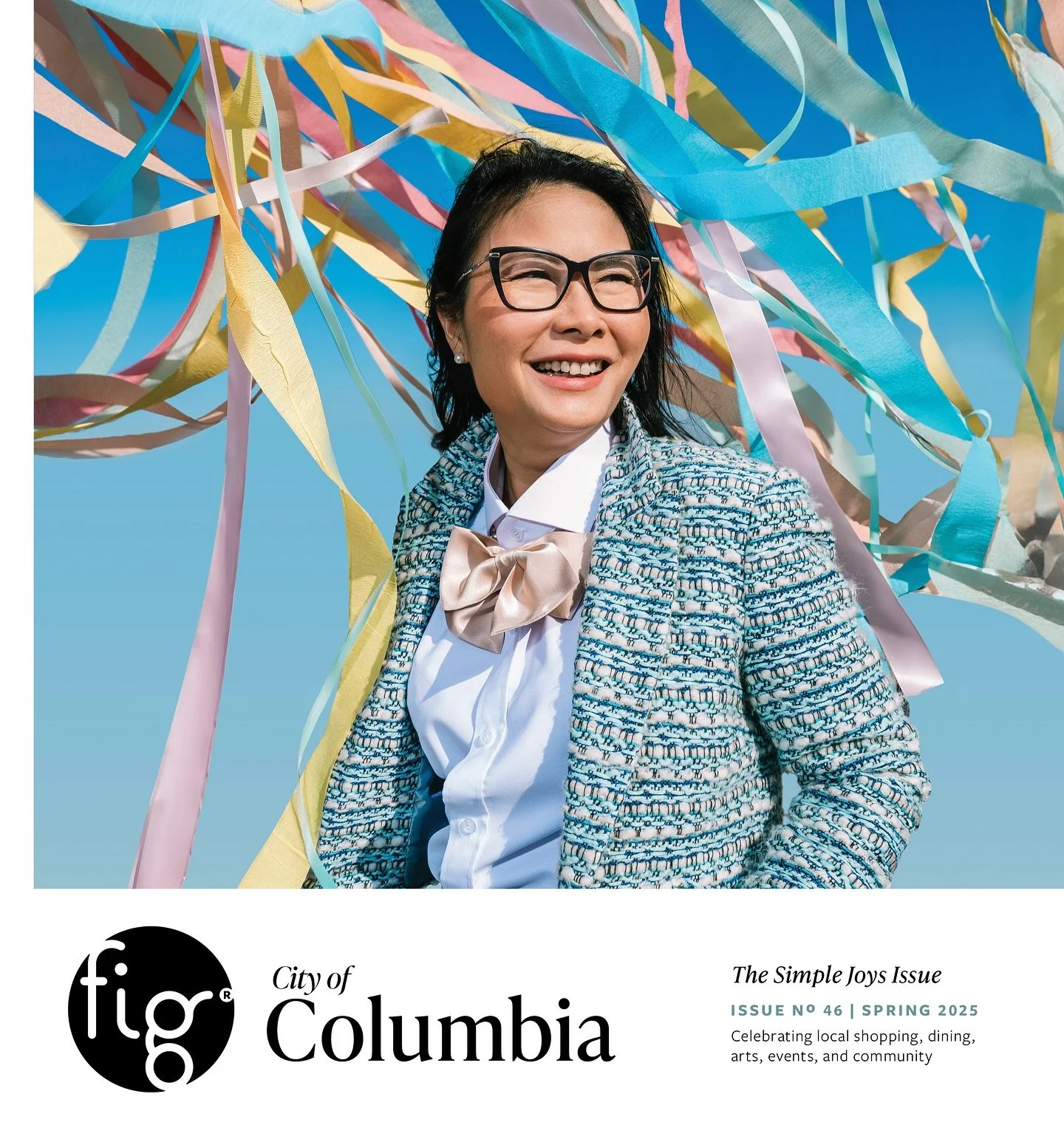 A woman wearing glasses and a patterned jacket with a bow tie, smiling outdoors in front of colorful streamers and a blue sky.