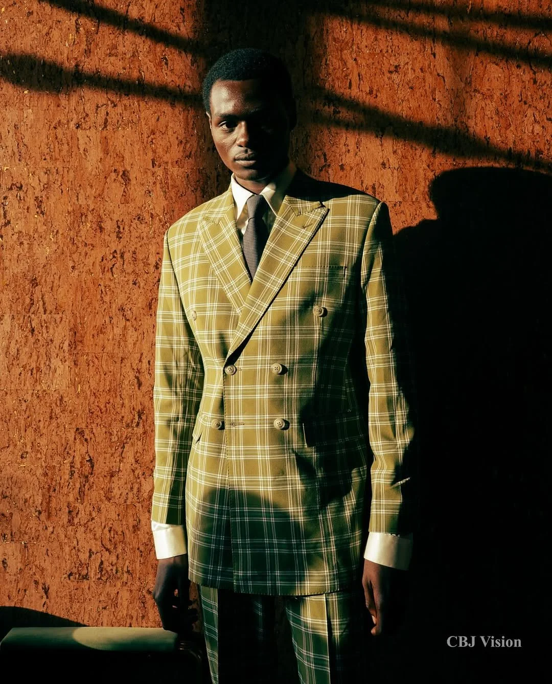 A young man in a yellow plaid suit, white shirt, and dark tie standing against a textured orange wall with shadows cast across his face and body.