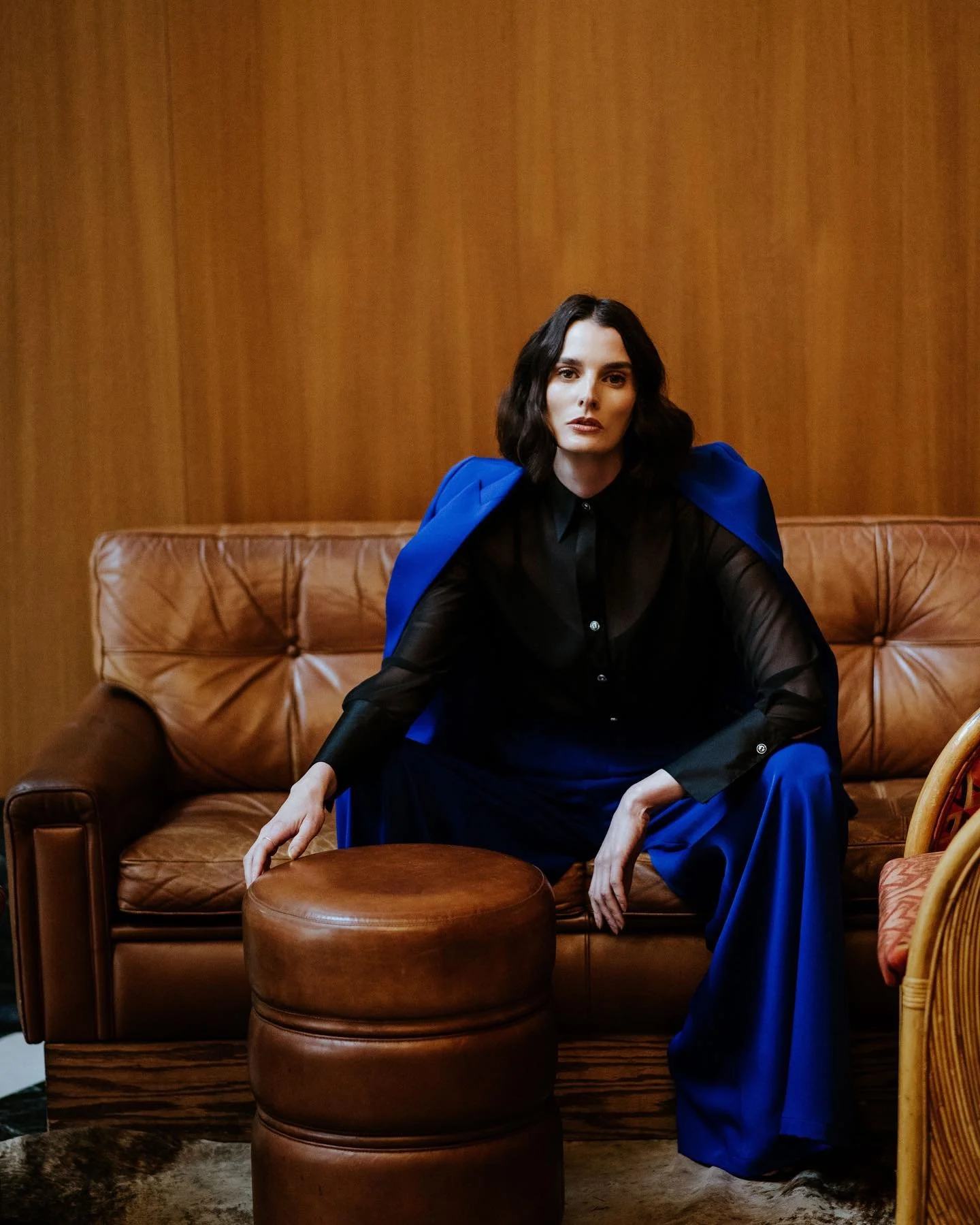 A woman with dark, wavy hair, wearing a black sheer blouse and bright blue pants, sitting on a brown leather sofa with a wood-paneled wall in the background.