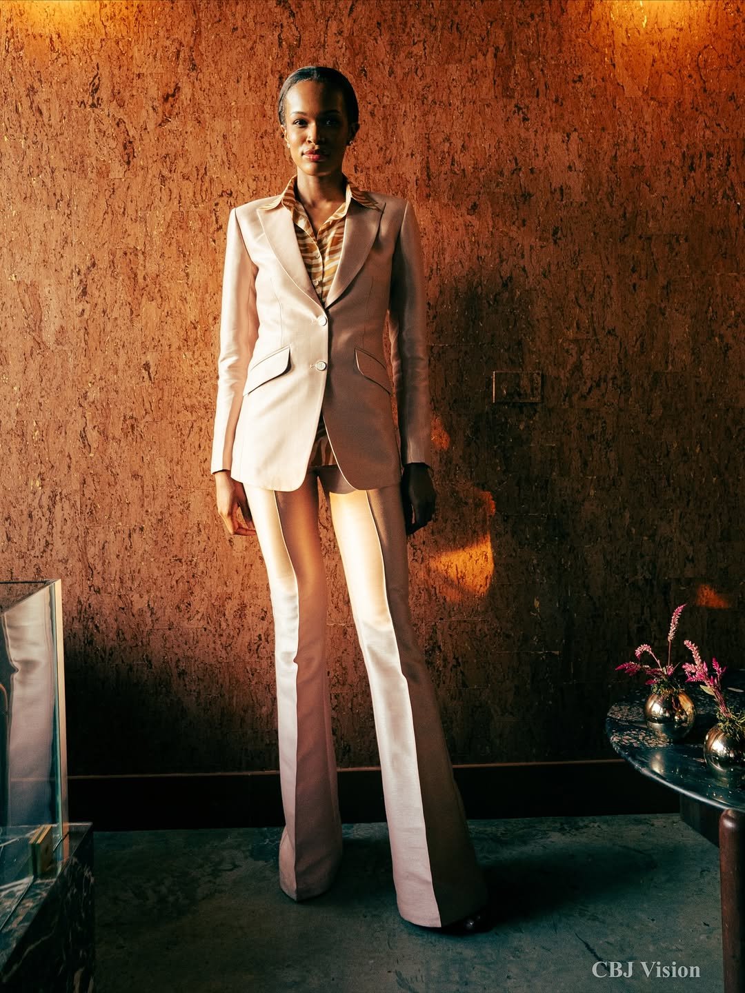 A woman wearing a beige blazer and matching wide-leg pants standing against a textured brown wall, with orange lighting on her left side.